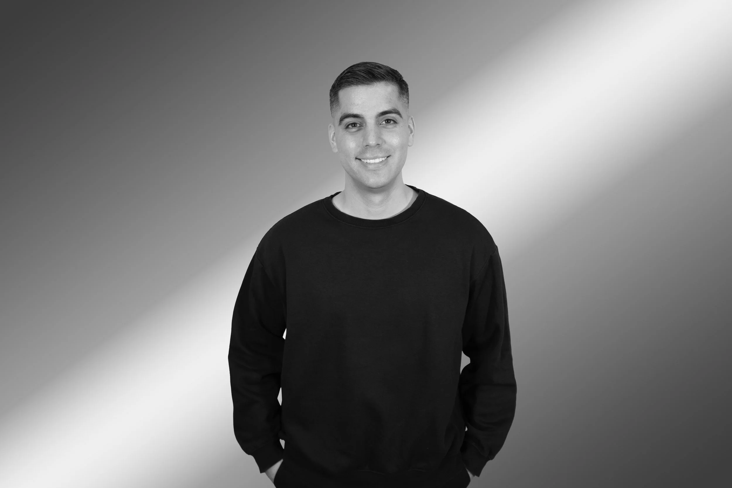 Black and white portrait of a smiling young man with short hair, wearing a dark sweatshirt, standing against a gradient background.