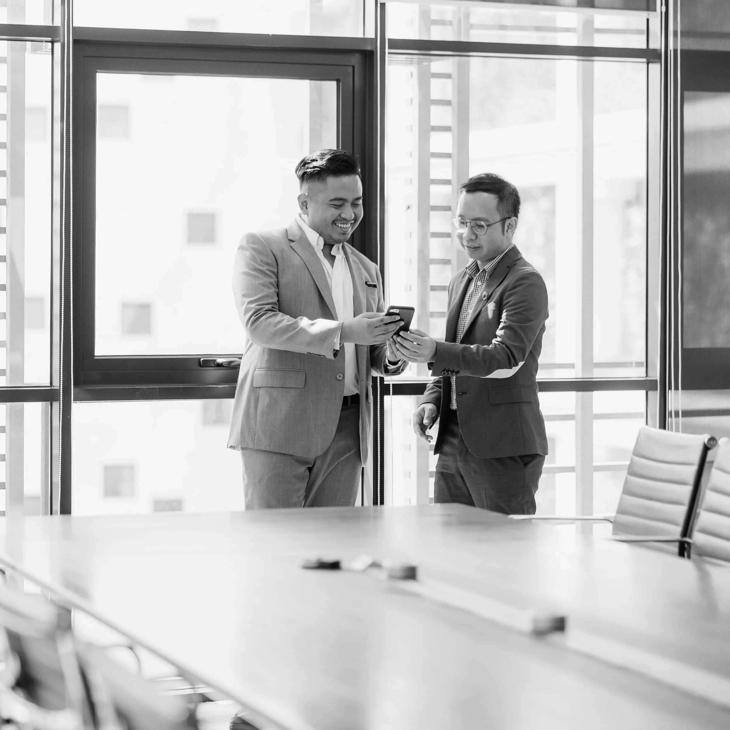 Two men in business suits sharing a moment while looking at a phone in a modern office conference room.