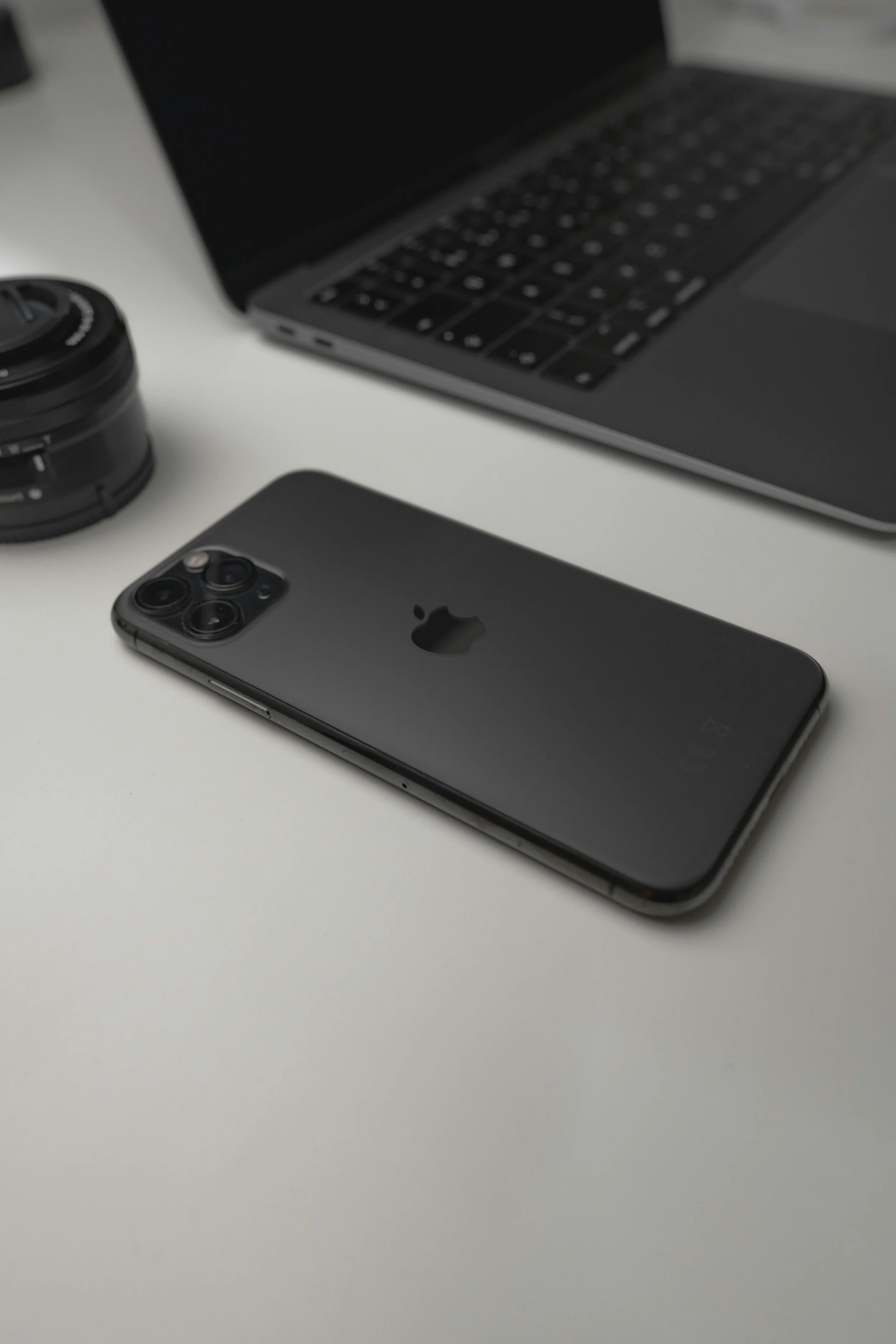 A black iPhone 11 Pro, a MacBook laptop, and camera lenses on a white surface.