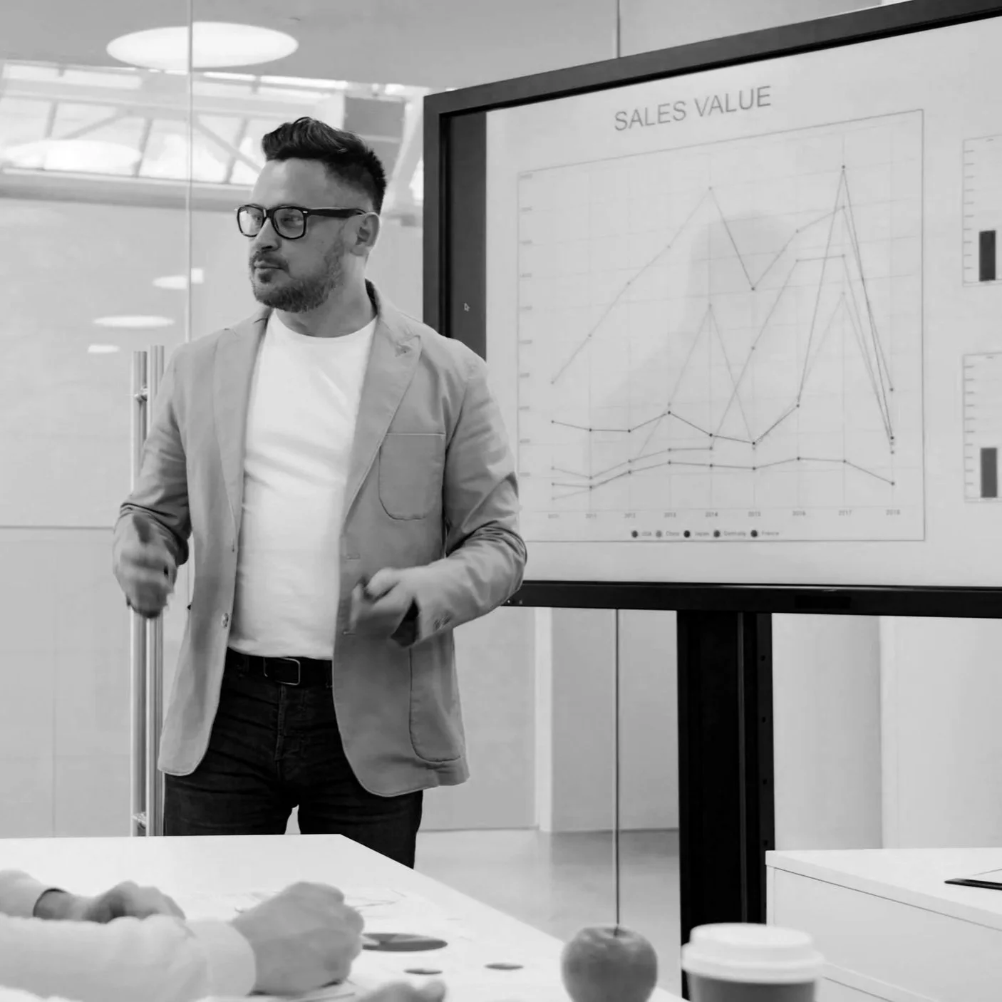 A man wearing glasses and a light-colored blazer giving a presentation in a modern office. Behind him is a screen displaying a line graph titled 'Sales Value', showing multiple fluctuating data lines across years. A person at the table is taking notes, with a coffee cup and an apple in the foreground.