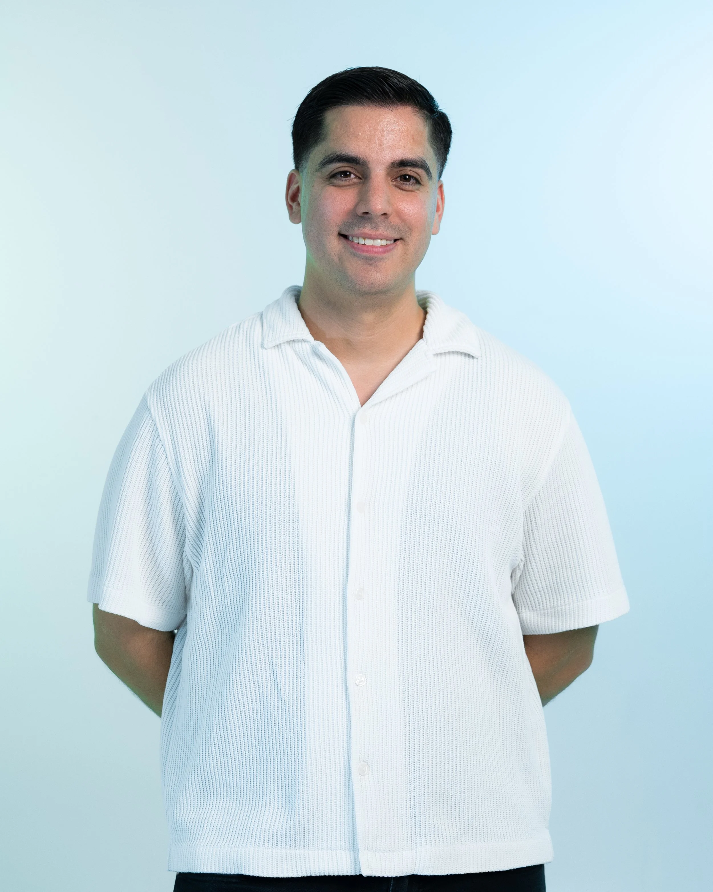 A man smiling, standing against a light-colored background, wearing a short-sleeved white polo shirt.