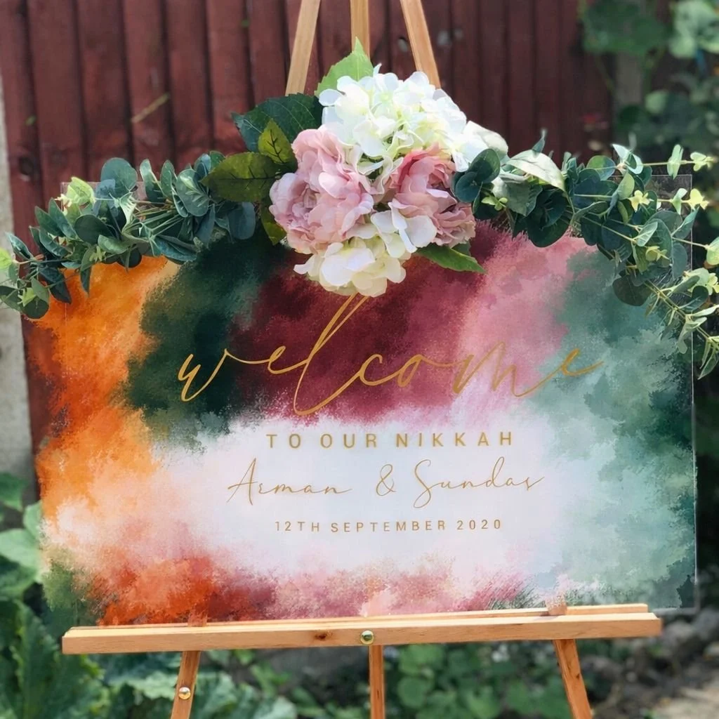 Welcoming sign with floral decoration for a wedding, featuring watercolor background with green, pink, and orange hues, and gold text that says "welcome to our Nikkah Aman & Sunday 12th September 2020".
