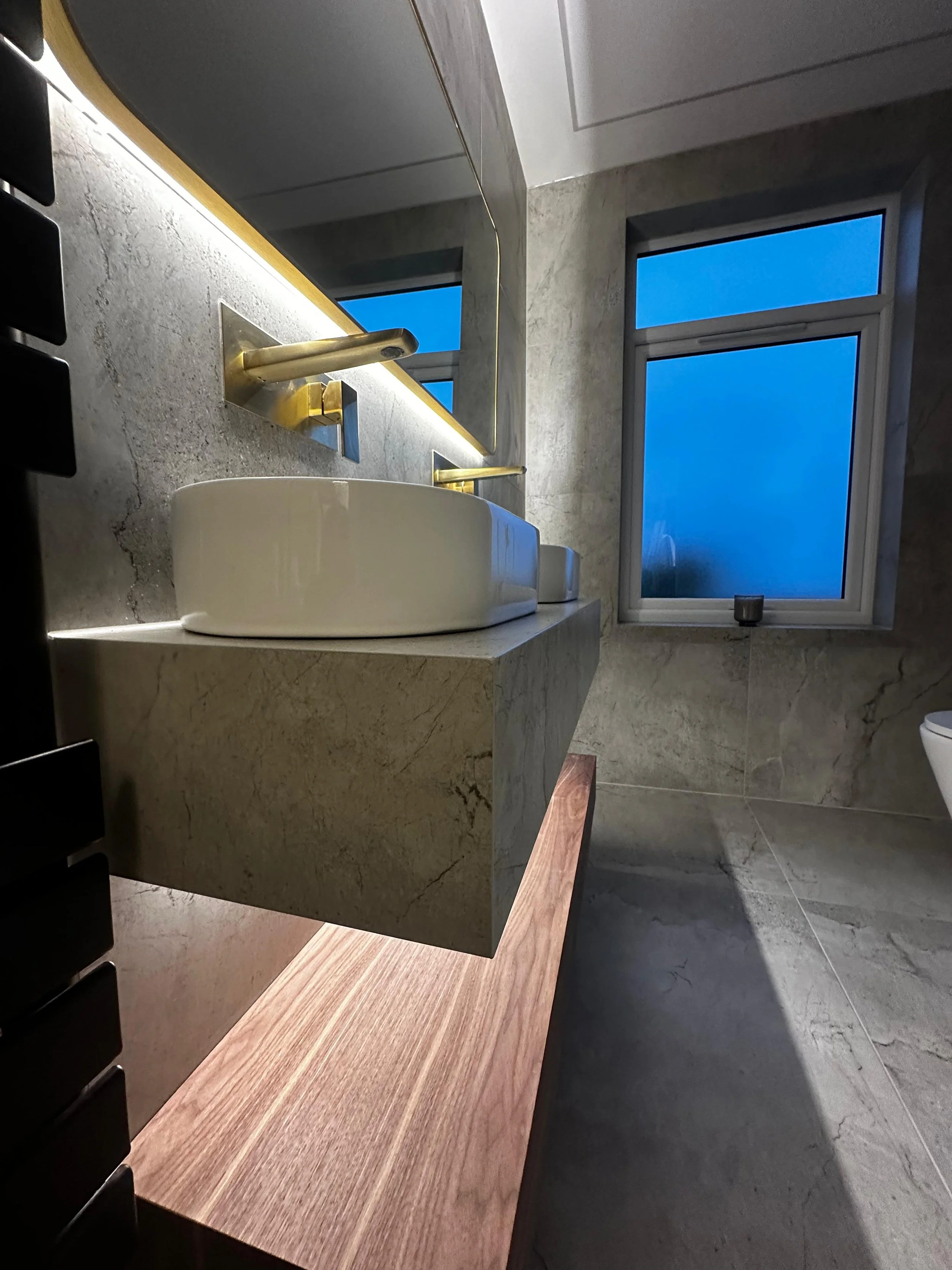 Modern bathroom with a vessel sink, gold faucet, large mirror, gray marble walls, and a window with a blue sky view.