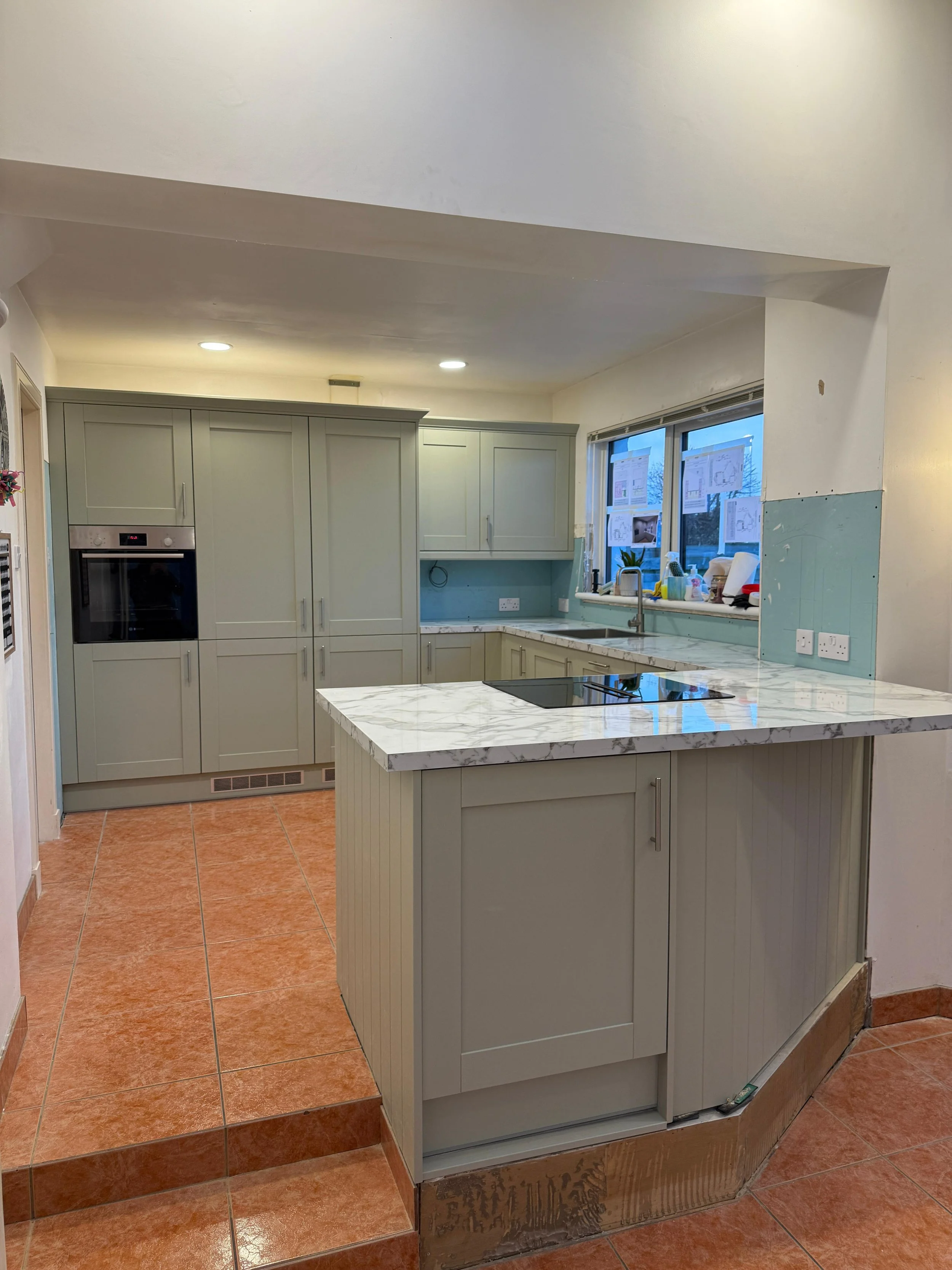 Modern kitchen with light gray cabinets, marble countertop island, terracotta floor tiles, large window, and various kitchen items on the windowsill.