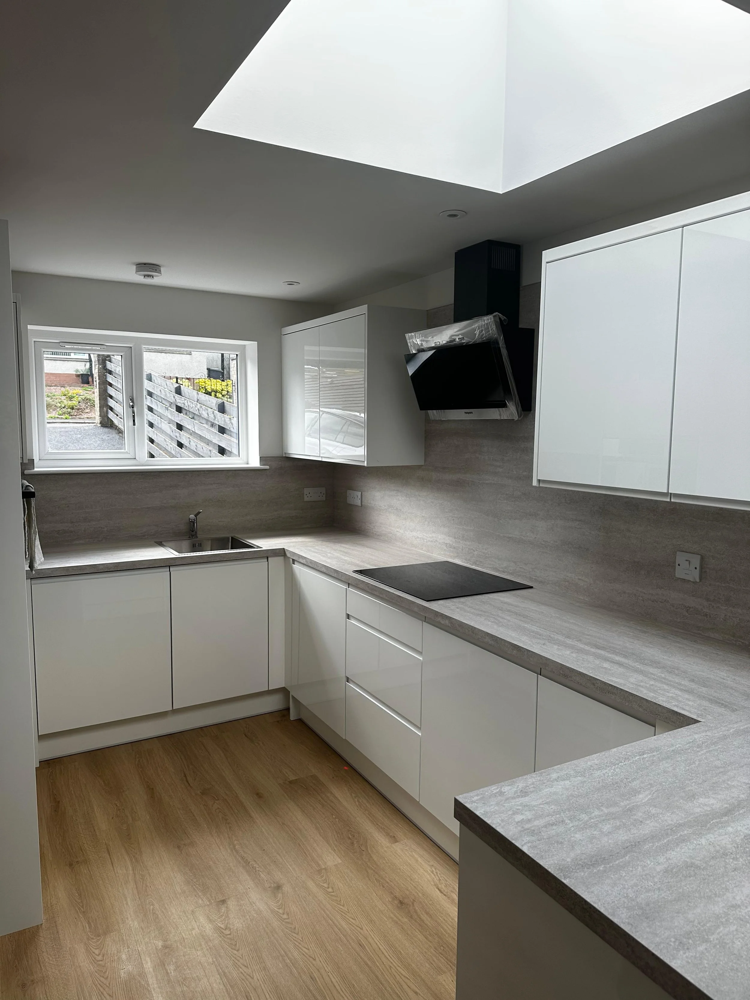 Modern white kitchen with wooden floor, a window above the sink, cabinetry, and a black stovetop, with natural light coming through skylight and window.