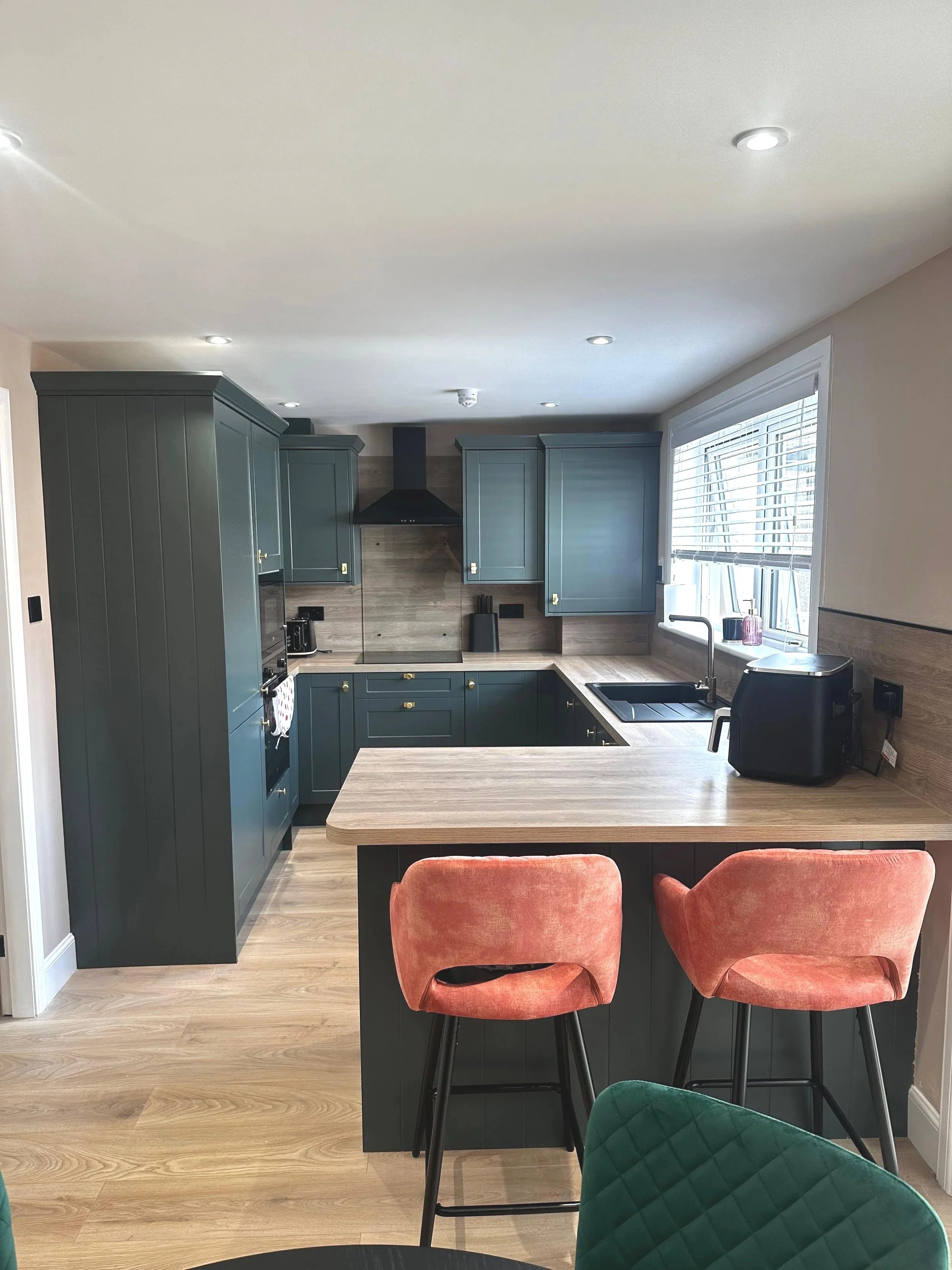 Modern kitchen with green cabinetry, wooden countertops, a window, and coral-colored bar stools.