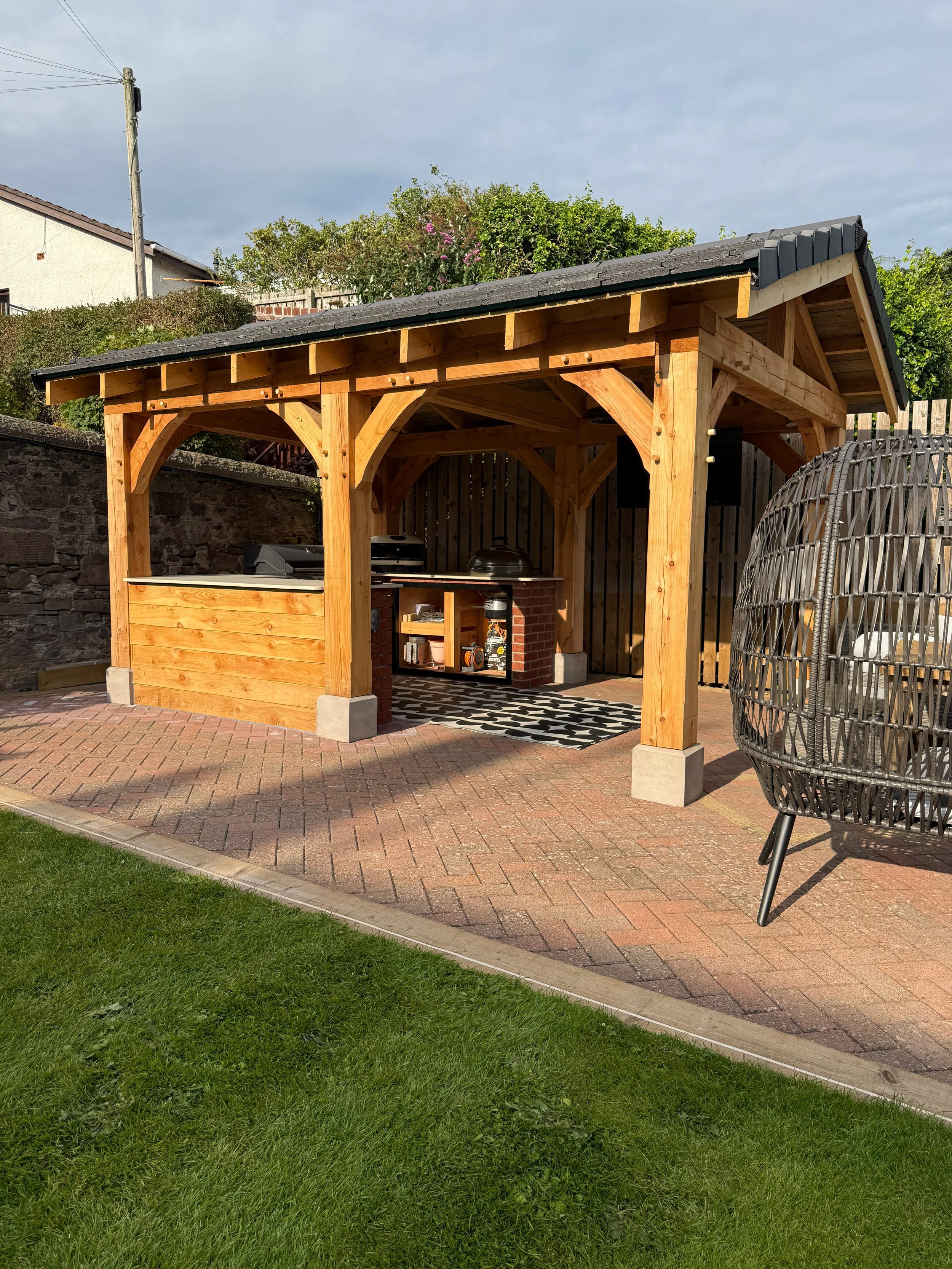 Wooden outdoor gazebo with a roof, brick and wood countertop, black and gray grills, situated on brick paving with a grassy lawn in the foreground.