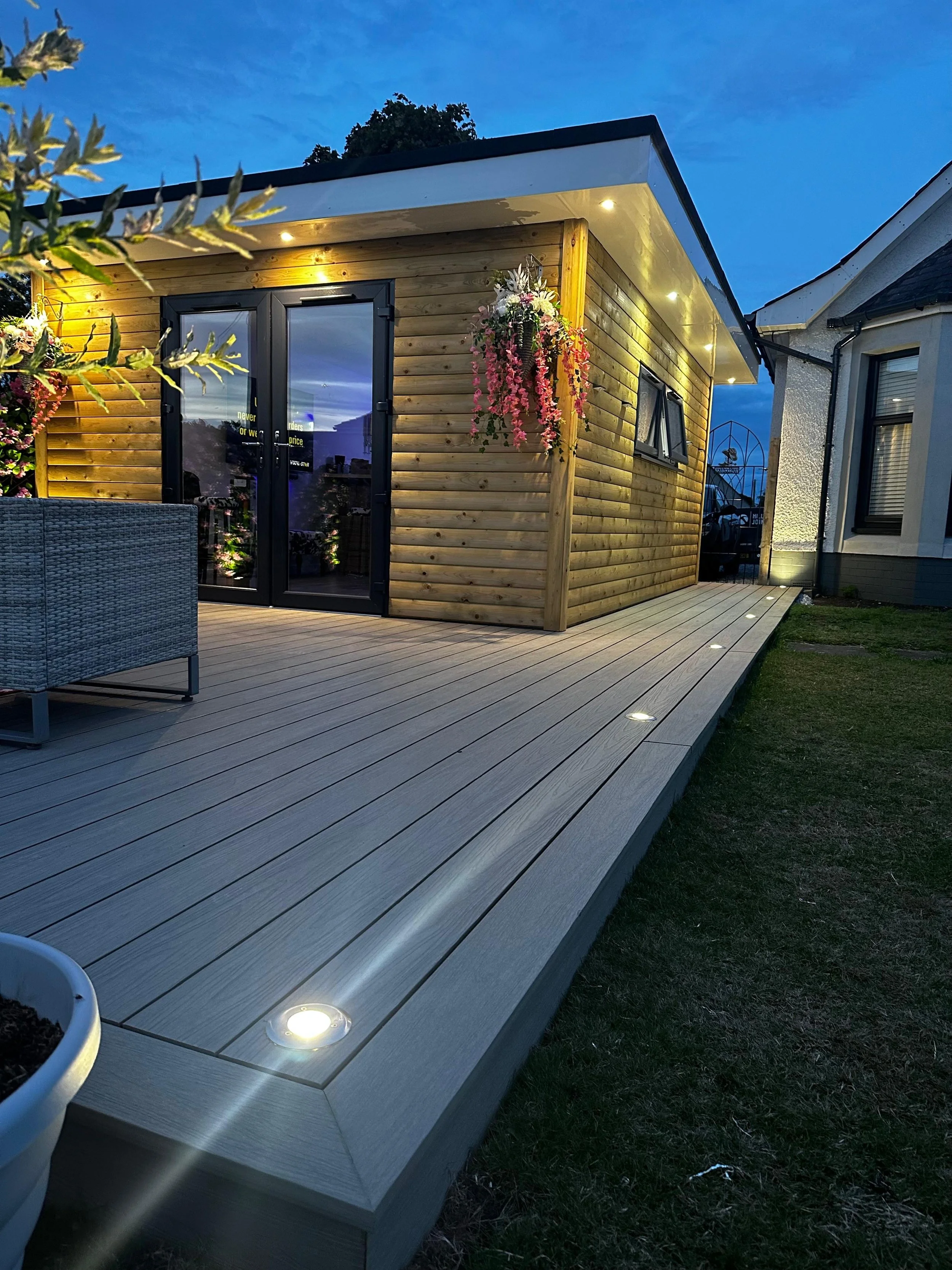 A tiny wooden house with glass double doors, illuminated outdoor lights, and hanging flower baskets, situated on a wooden deck at dusk.