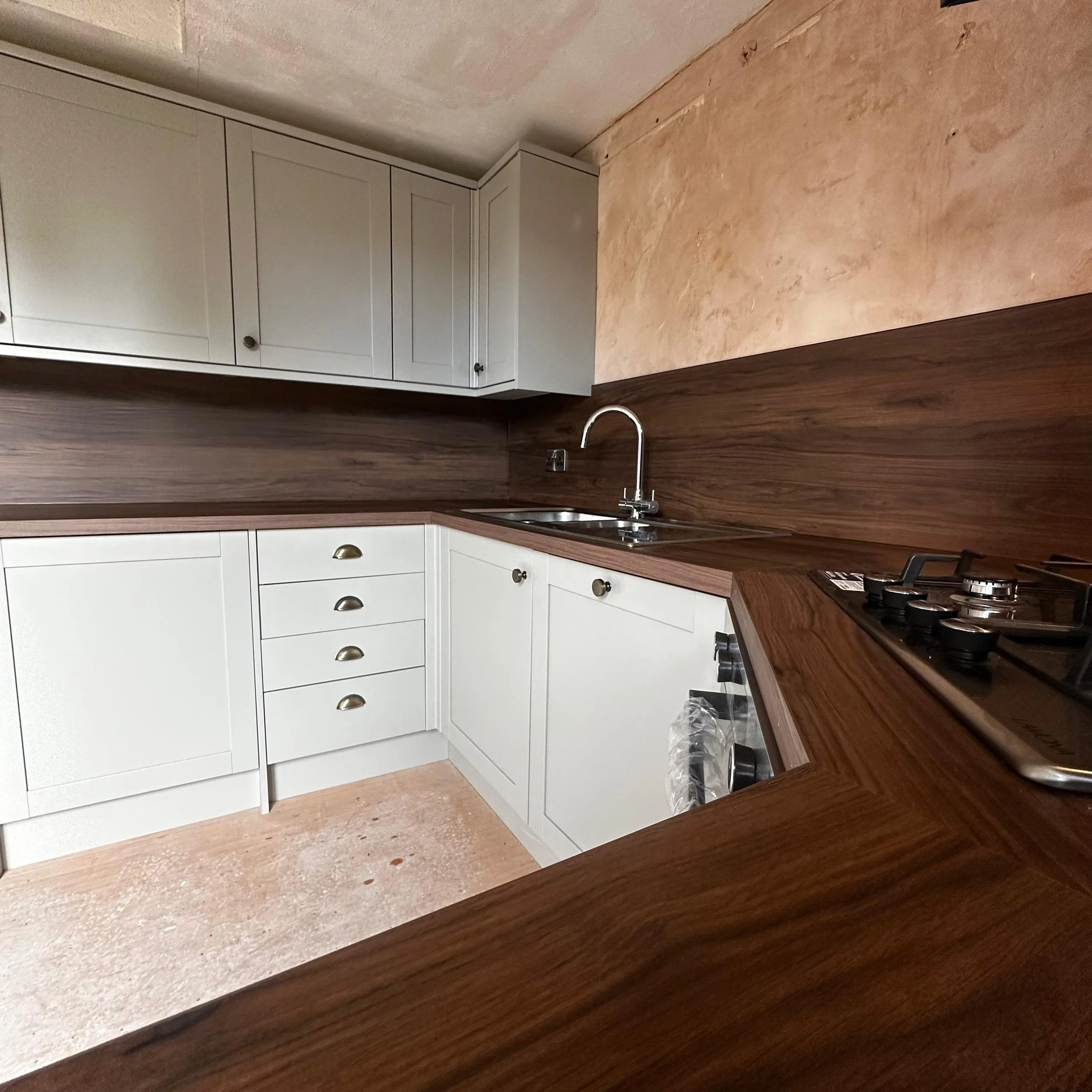 A modern U-shaped kitchen featuring white cabinets, a wooden countertop and backsplash, a stainless steel sink with faucet, and a gas stove, with a textured peach-colored wall and a wooden floor.