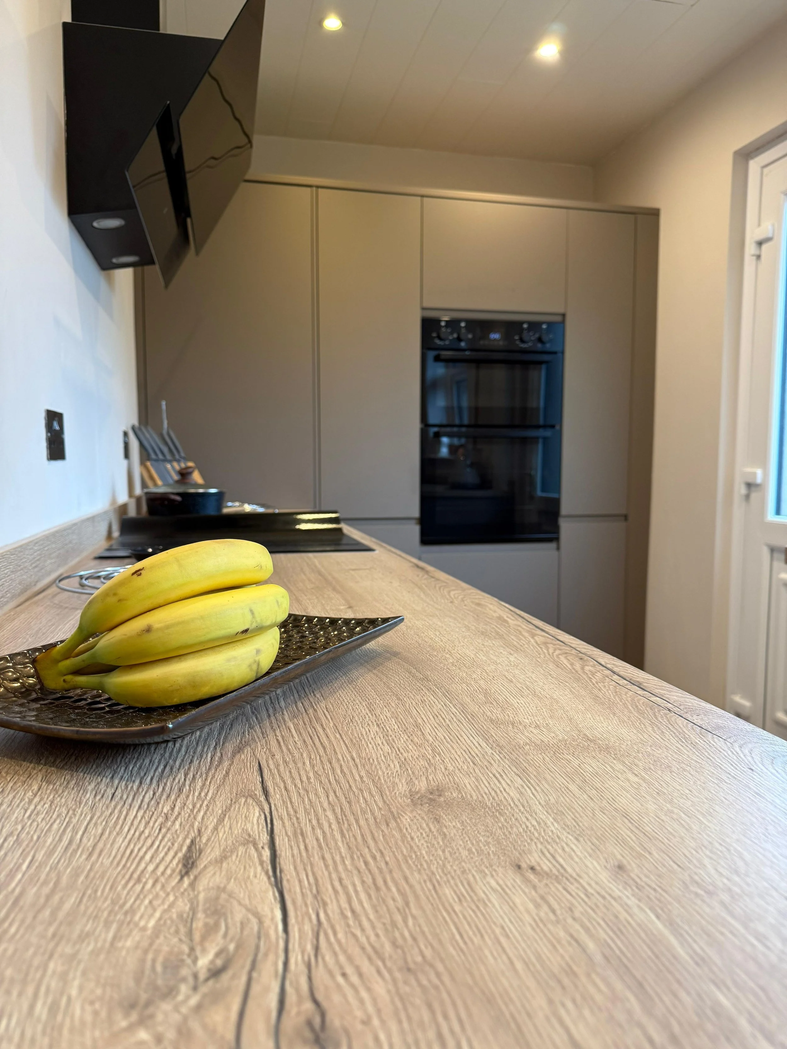 A kitchen with a wooden countertop, a bunch of bananas on a decorative plate, a wall-mounted television, and built-in oven, with ceiling lights.