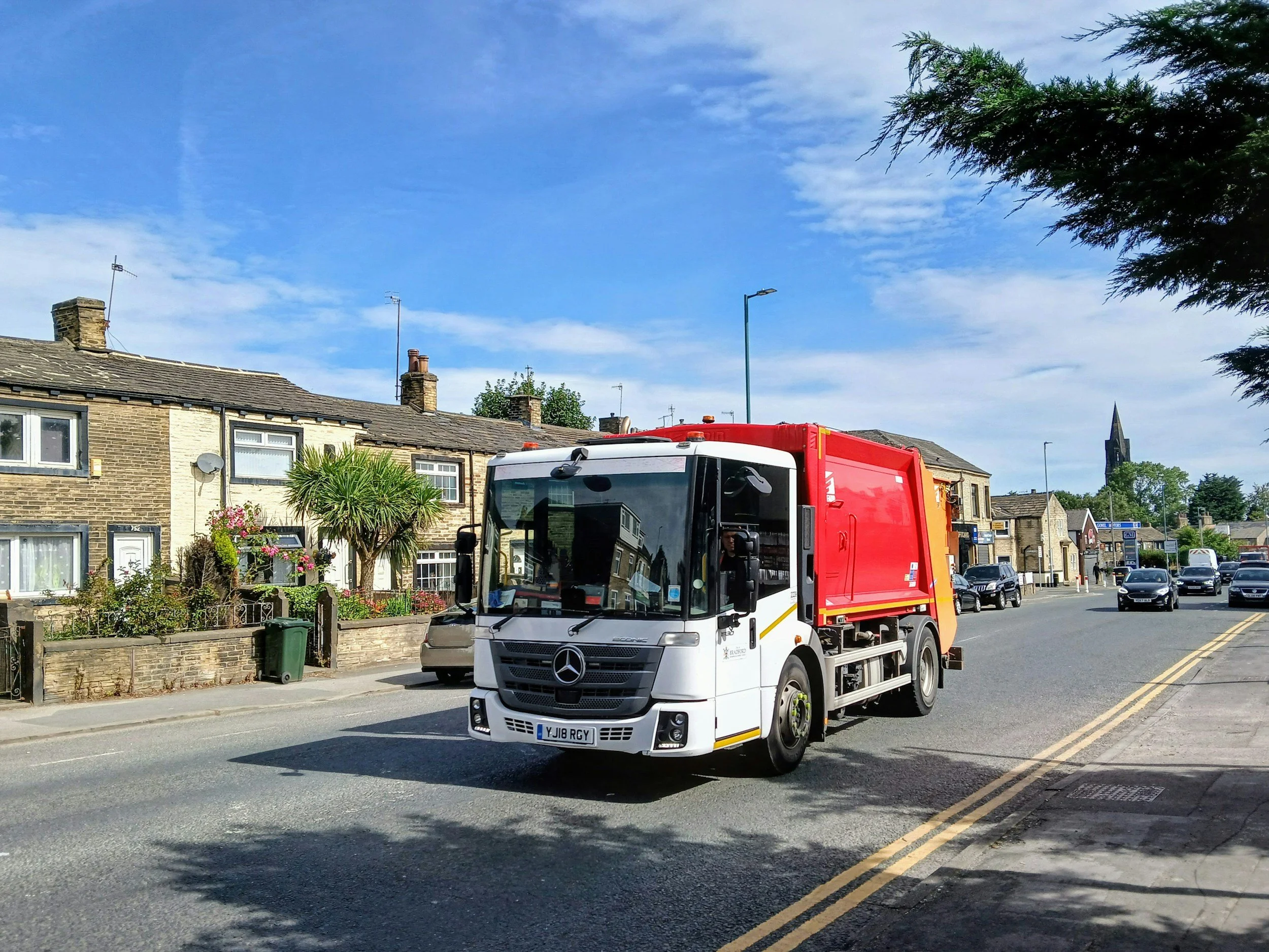 White and red Mercedes-Benz street sweeper truck driving on a city street with residential buildings and a church steeple in the background under a partly cloudy sky.