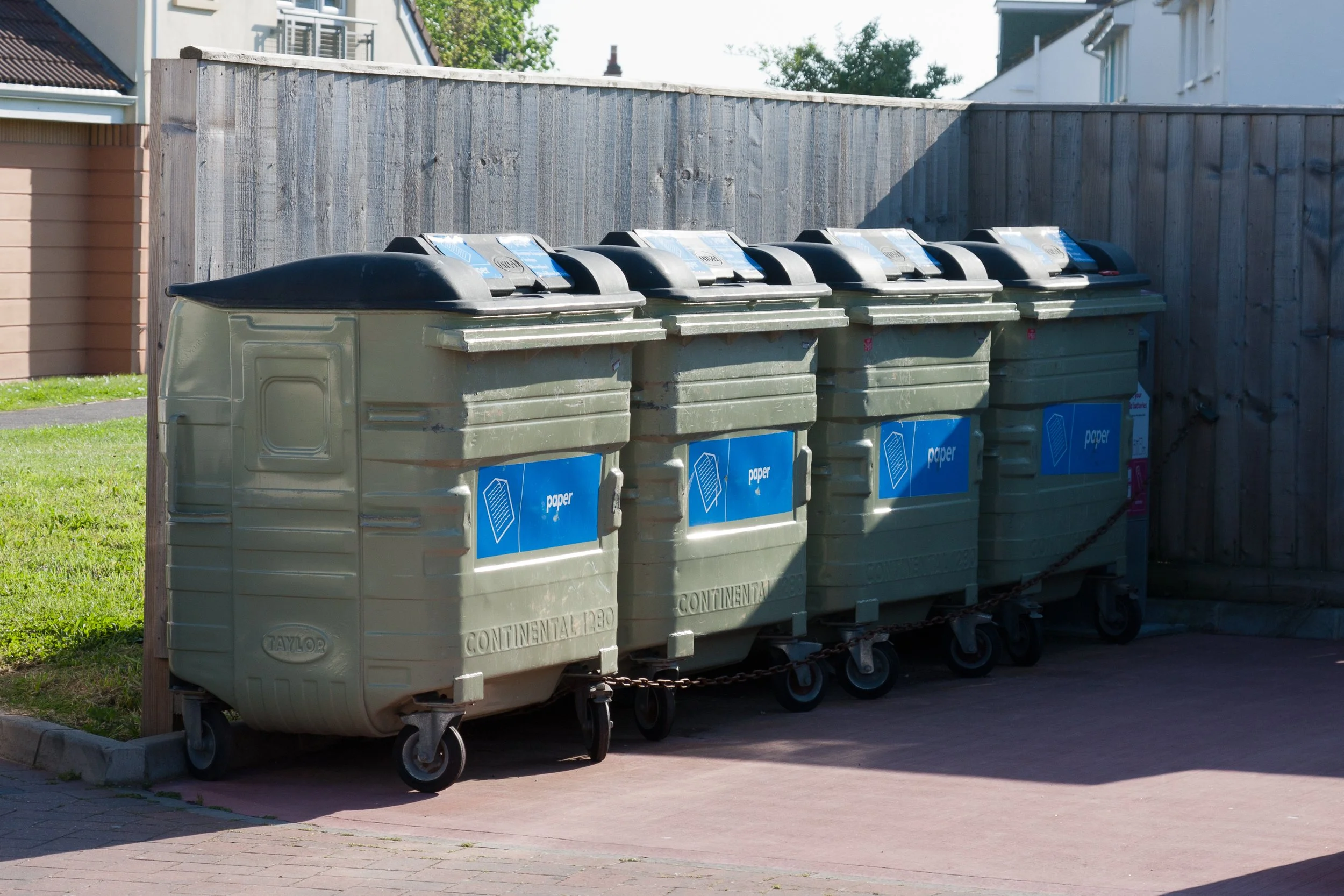 Four green waste and recycling bins with black lids lined up outside on a paved surface, with a wooden fence behind them and residential houses in the background.
