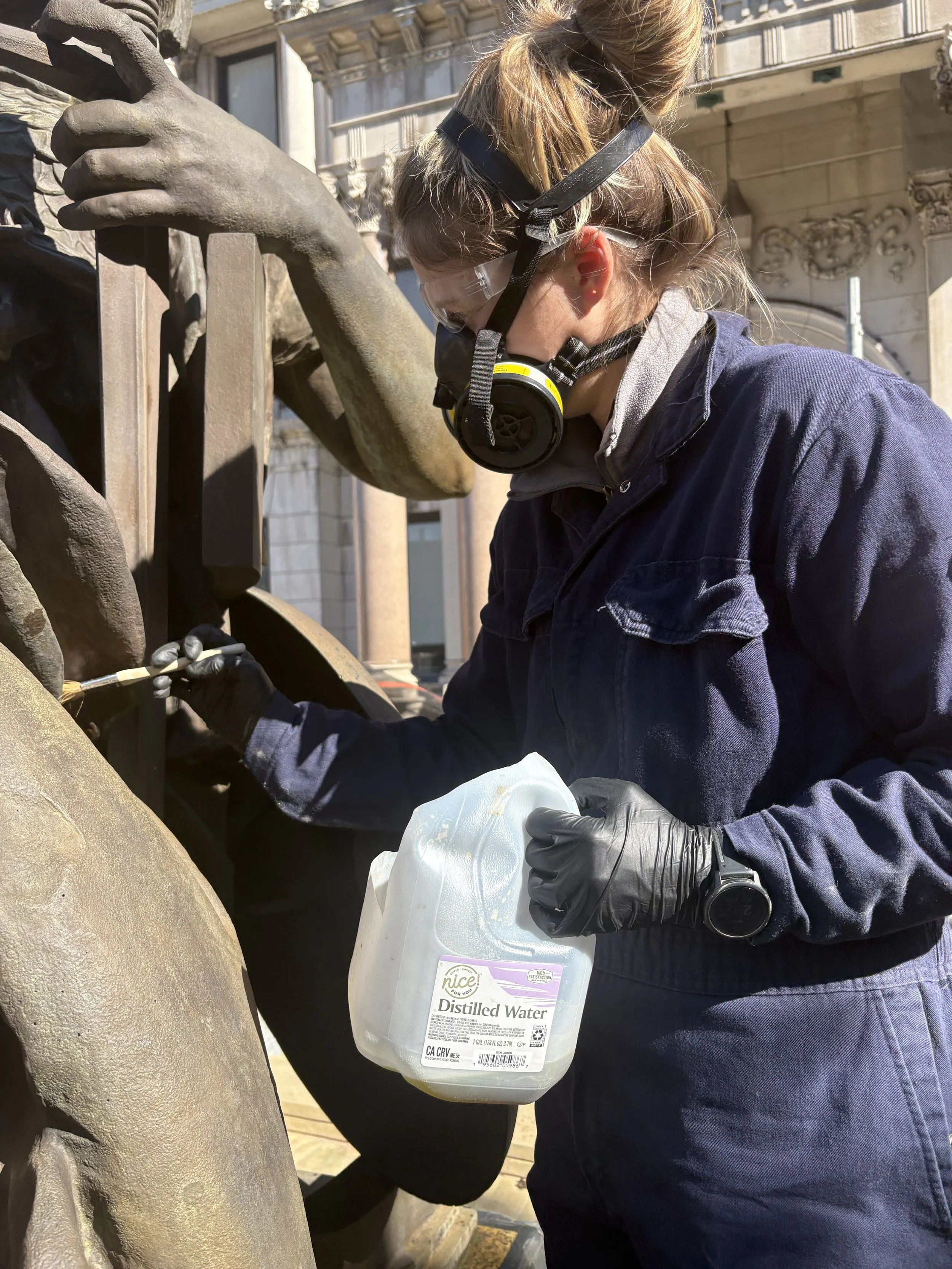 Person wearing safety glasses and face mask, holding a quart-sized container of distilled water, cleaning or restoring a bronze or metal sculpture outdoors.