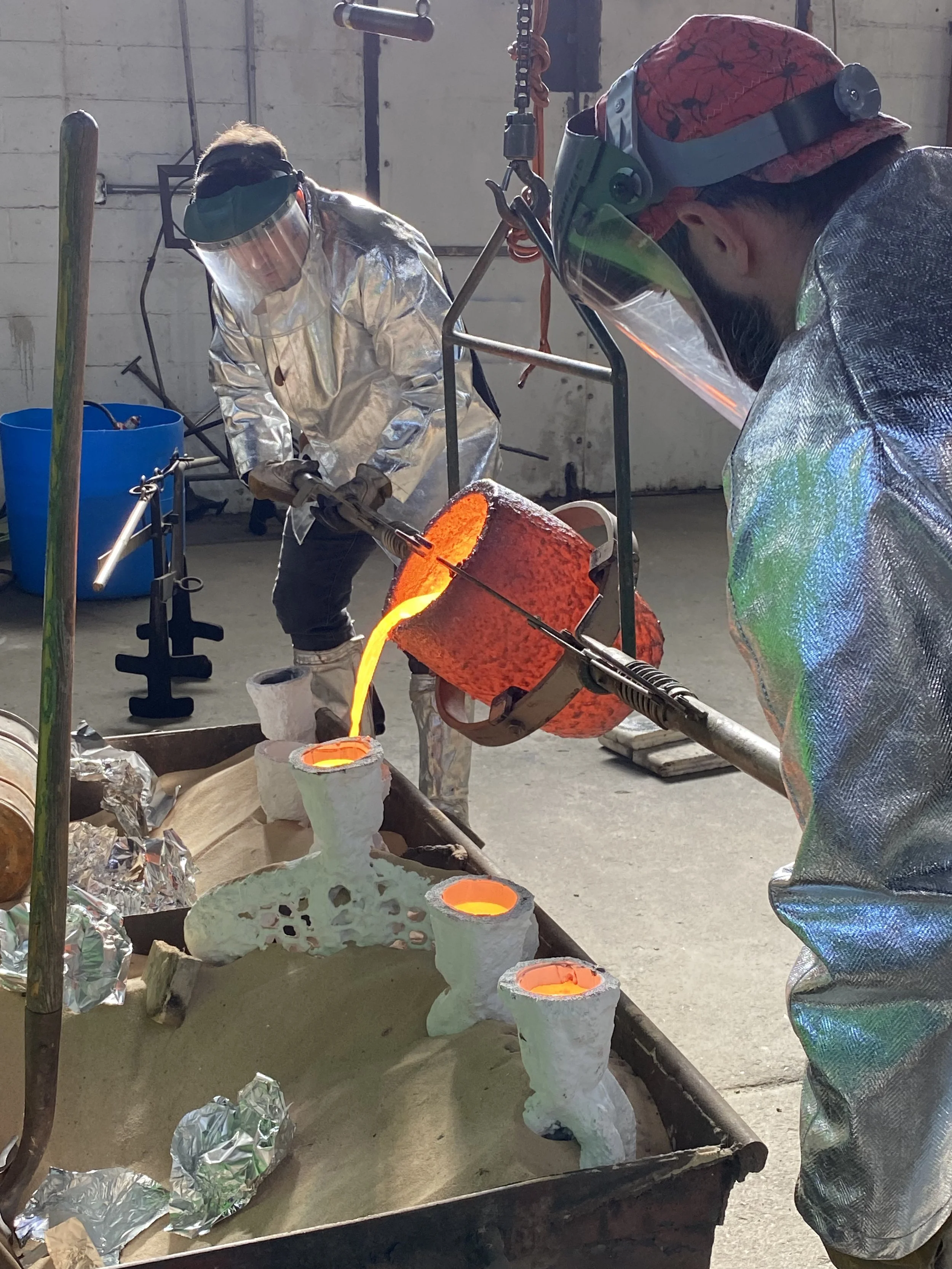 Two glassblowers wearing protective gear, including face shields and reflective aprons, working with molten glass in a workshop. They are shaping or blowing glass art pieces, surrounded by tools and protective materials.