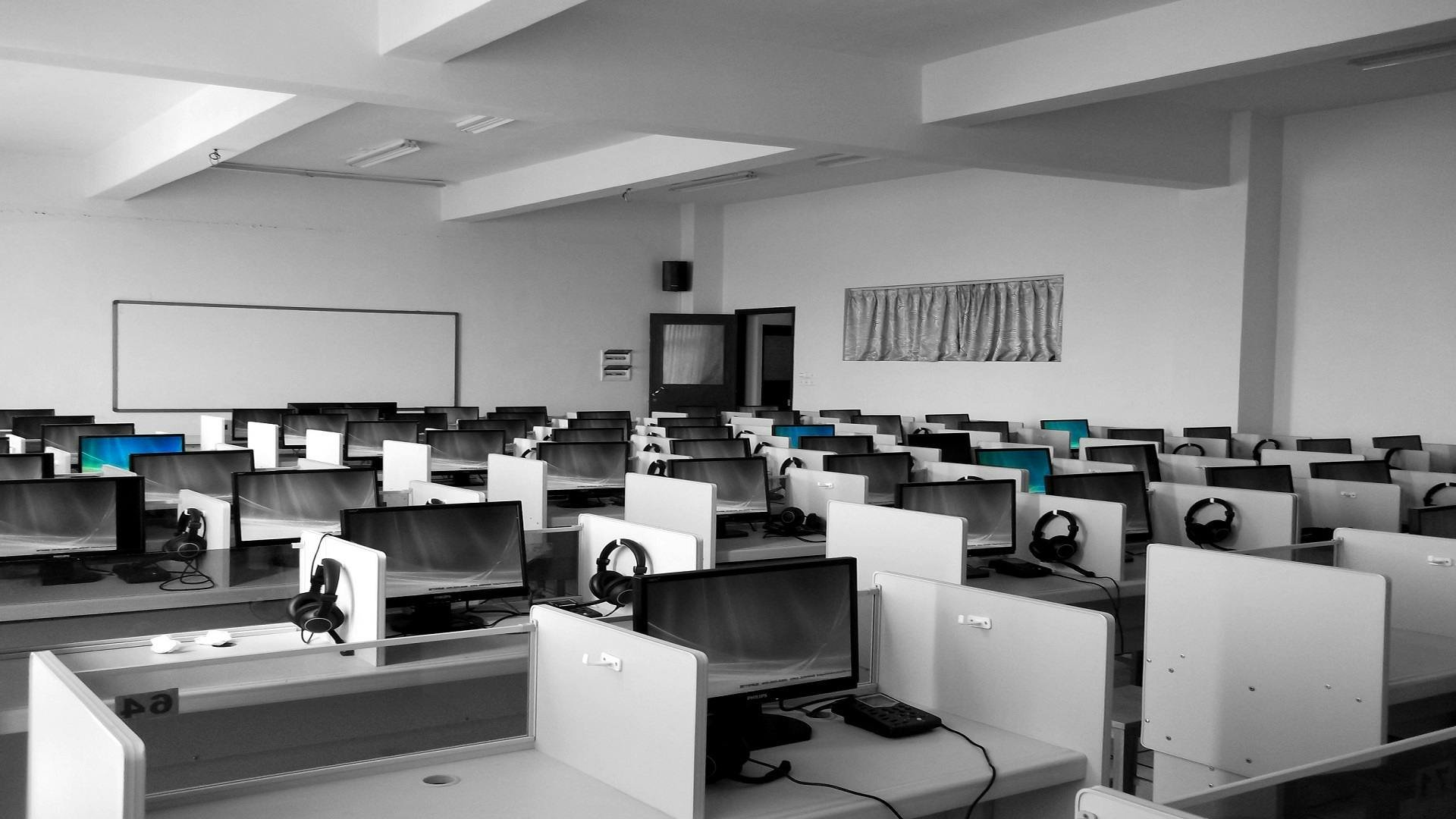 Empty computer lab with rows of desks and monitors, some with headsets, in a room with white walls, fluorescent lighting, and a window with curtains.