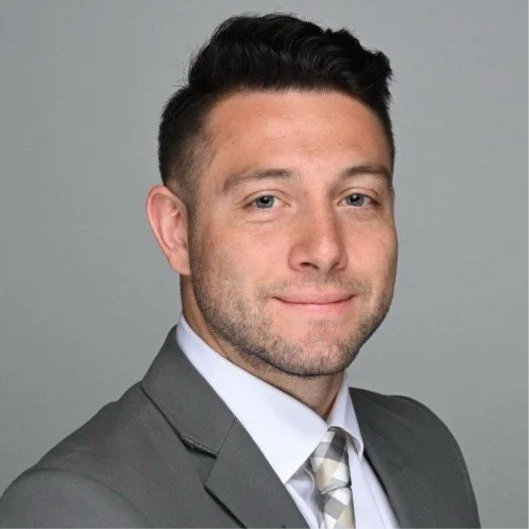Professional headshot of a young man in a gray suit, white shirt, and plaid tie, with short dark hair, smiling against a neutral gray background.