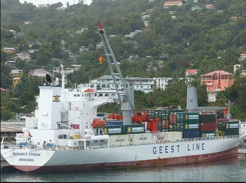 Cargo ship named Geest Line with containers, docked near a hillside with residential buildings.
