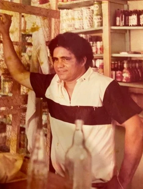 A man with curly hair and a light-colored shirt with dark horizontal stripes stands inside a store with shelves of products behind him. He is smiling and resting his arm on a wooden beam.