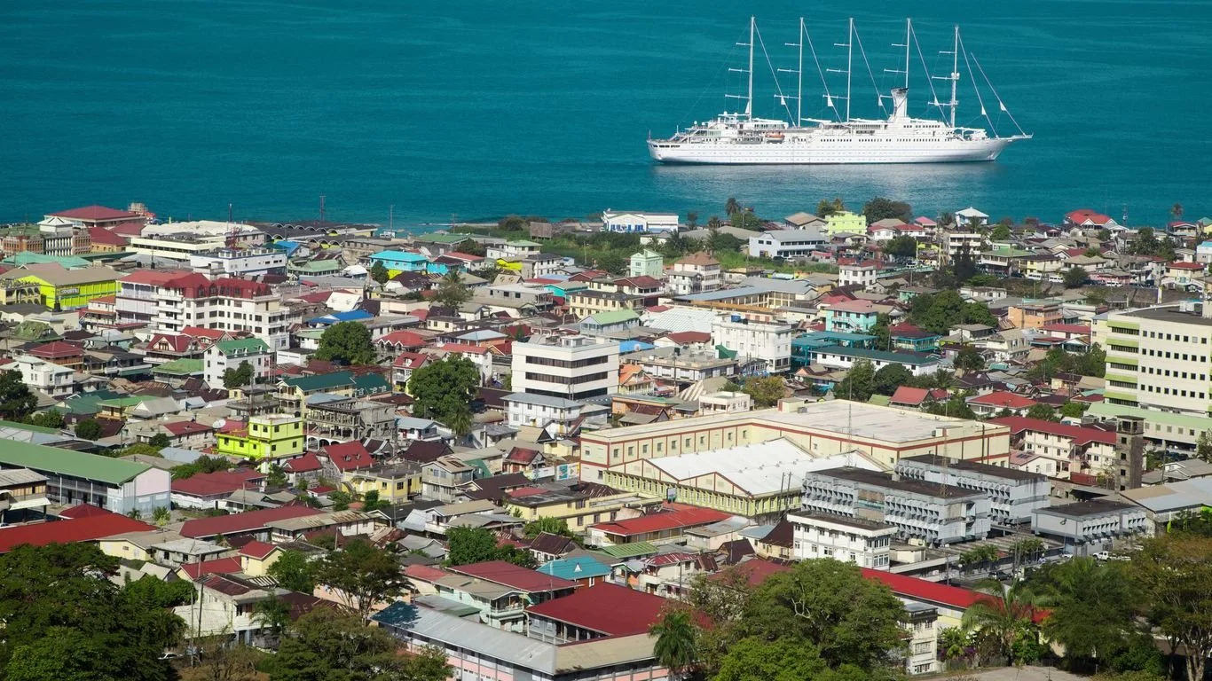 A coastal city with a variety of colorful buildings and houses, a large white sailboat sailing in the ocean, and lush green trees in the foreground.