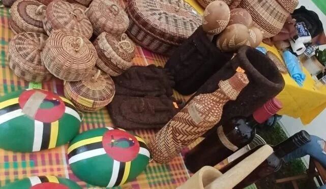Display of traditional woven hats, wooden and ceramic sculptures, colorful painted oval objects, and bottles, set on a checkered tablecloth.