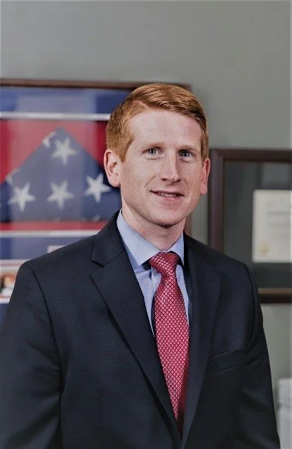 A young man with red hair in a suit and pink tie standing in an office or government building with American flags and framed documents in the background.