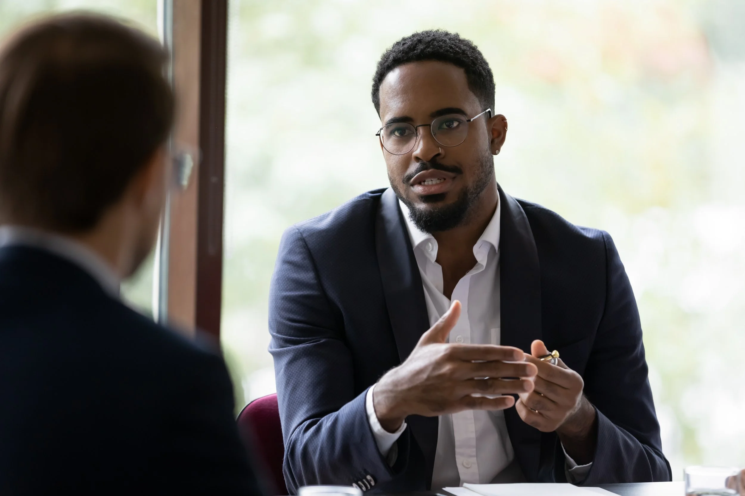 A man with glasses, a beard, and dark curly hair wearing a dark blazer and a white shirt, engaged in conversation with another person during a meeting or interview.