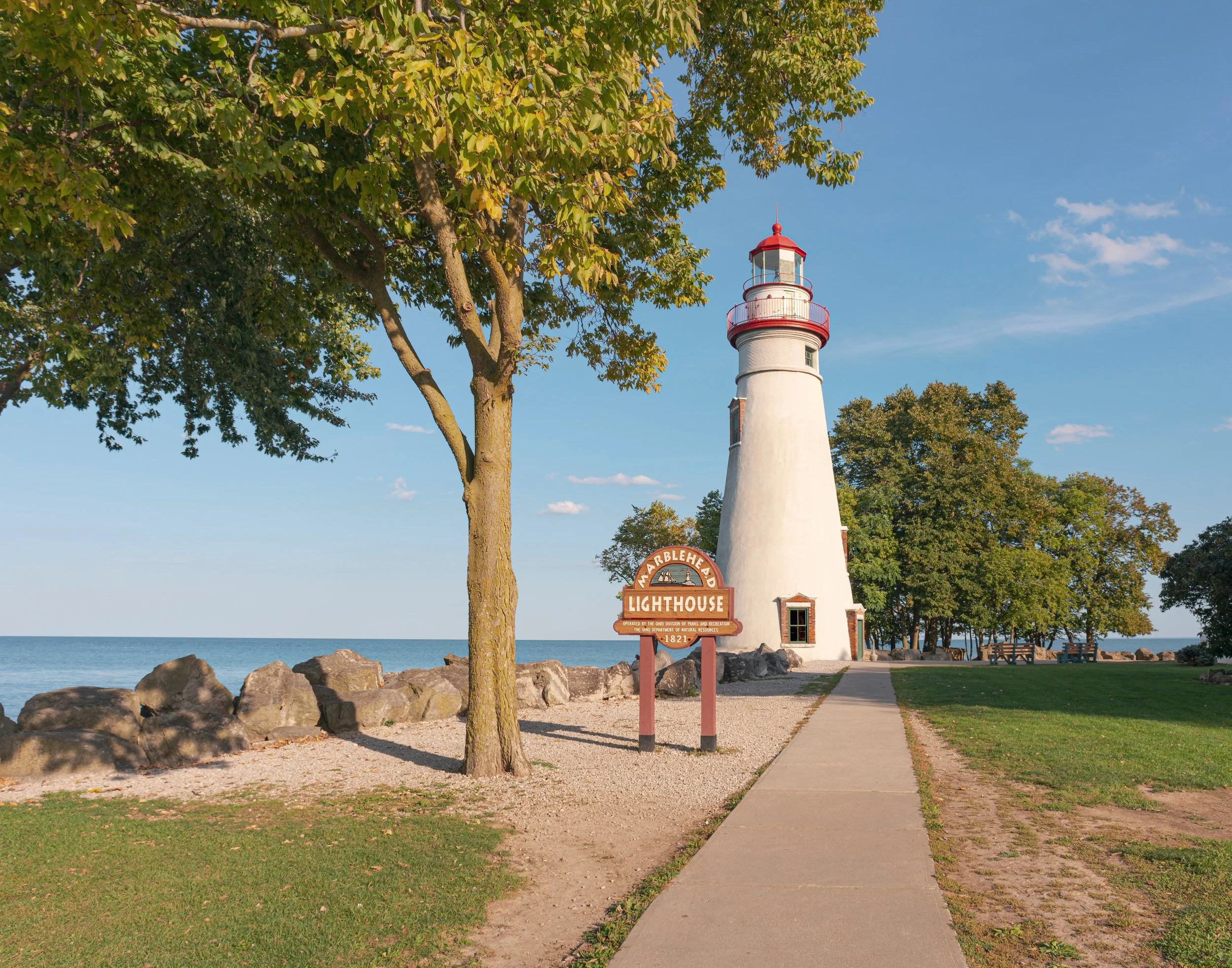 A lighthouse by a lakeside, with a sign reading 'Marblehead Lighthouse 1821', a tree, rocks, a sidewalk, and green grass under a clear blue sky.