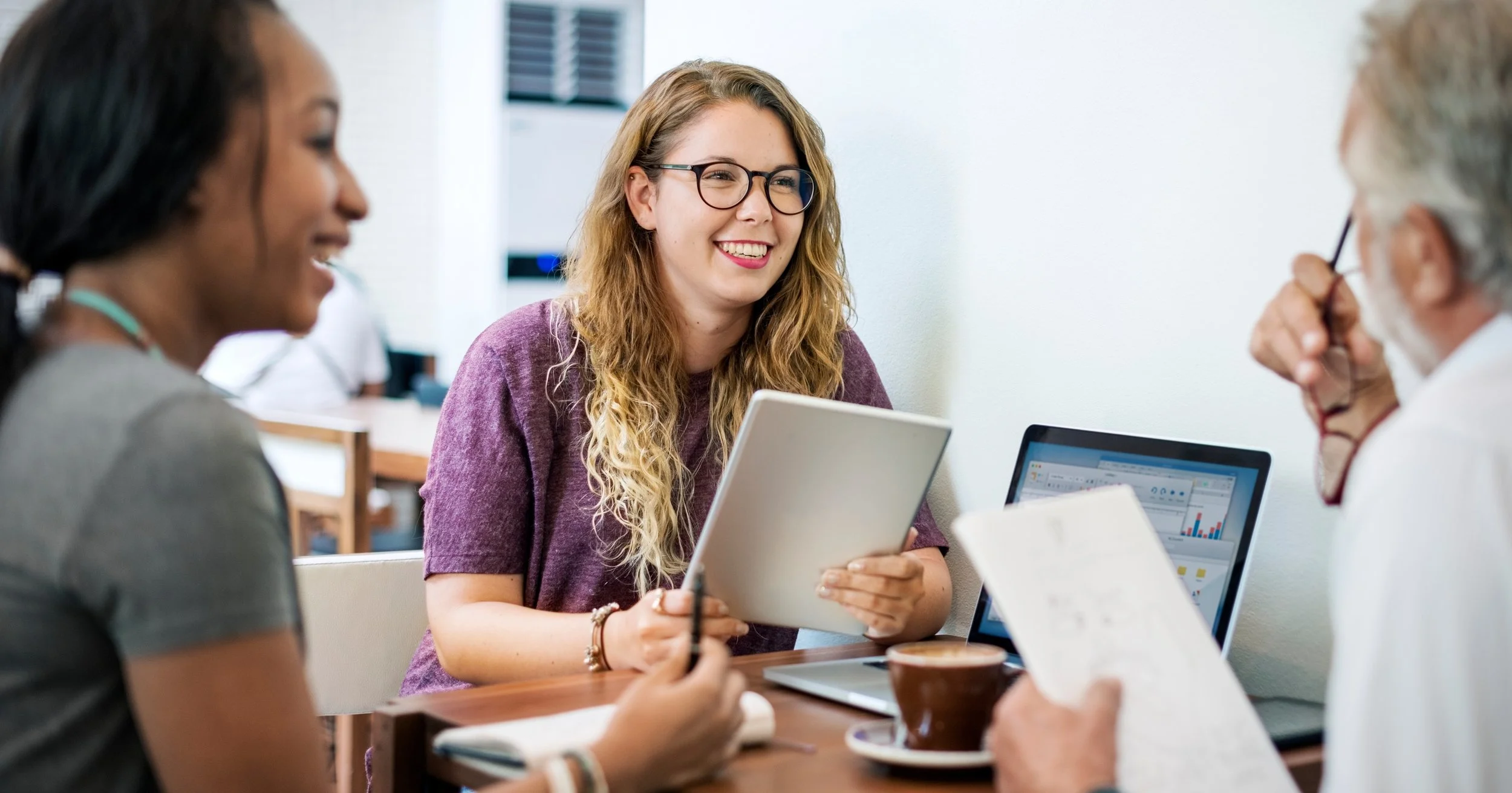 Three people in a meeting, a woman with glasses and curly hair smiling, holding a tablet, engaging with an older man with gray hair and glasses, and another woman, in a bright office setting.