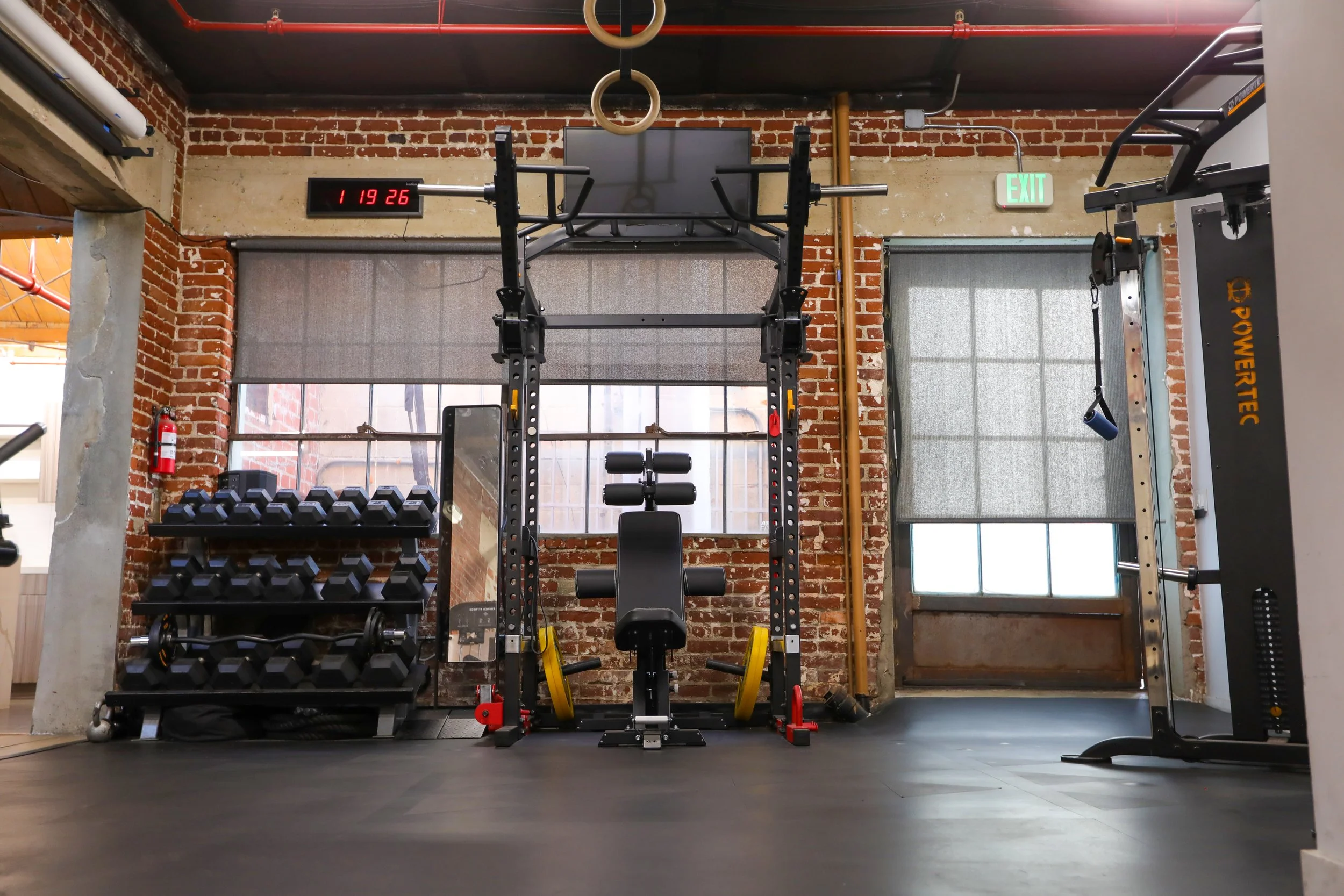 Symmetrical front view of private Los Angeles fitness studio with exposed brick walls, dumbbell rack, squat rack, and open floor space for personal training and content creation.
