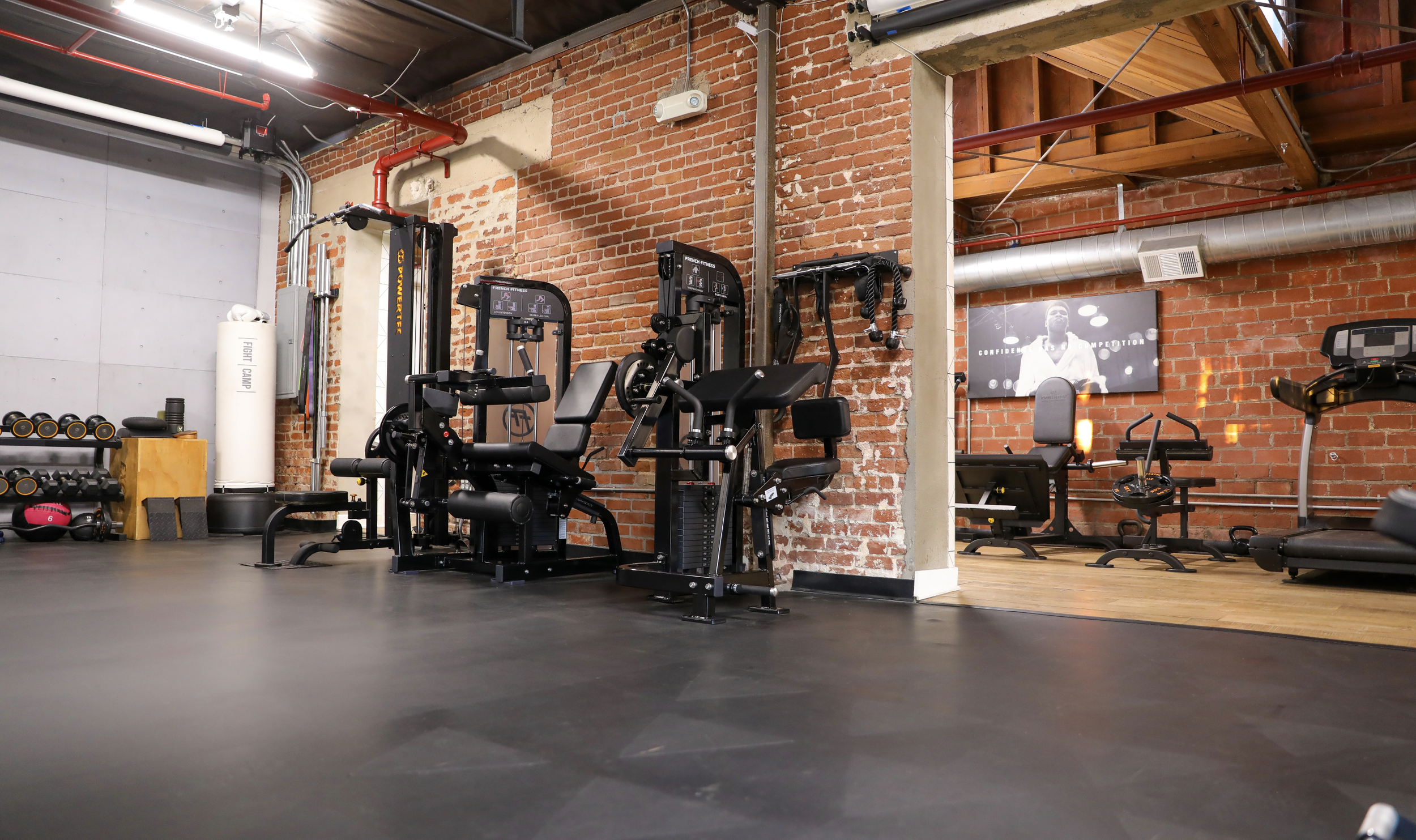 Fitness gym with various exercise equipment including machines, free weights, a punching bag, and a treadmill, with exposed brick walls and a monitor screen on the wall.