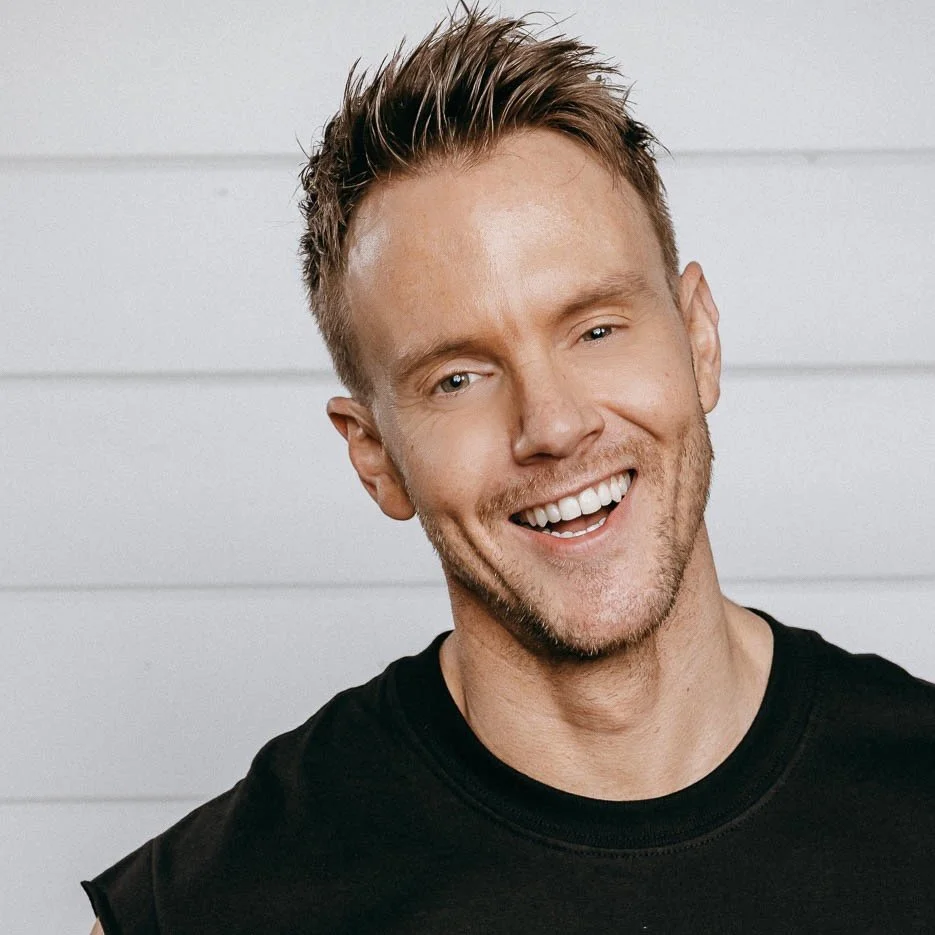 A smiling man with short, styled hair, light skin, and a beard, wearing a black t-shirt, standing against a white wooden background.