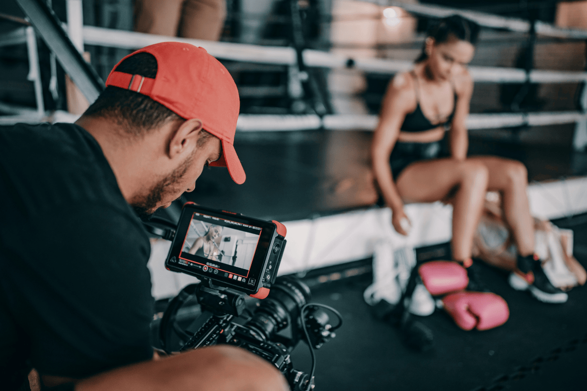 A man in a red cap filming a woman sitting on gym benches with workout equipment nearby.