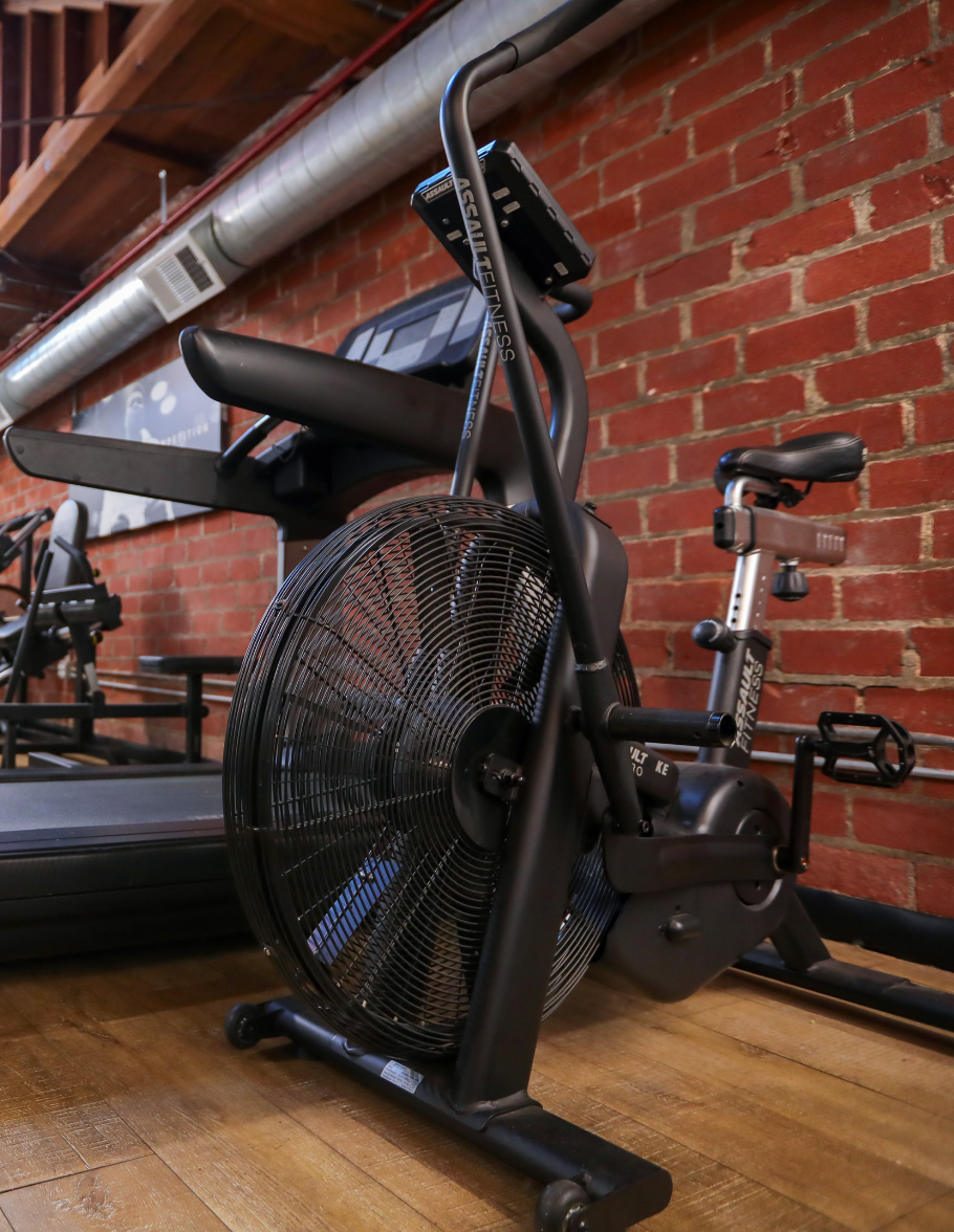 Black exercise bike with an oversized front fan, placed on a wooden floor, against an exposed brick wall in a gym.