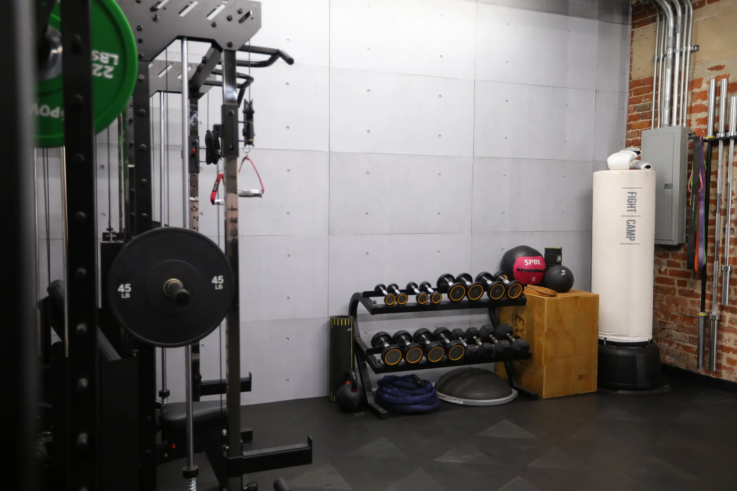 Gym corner with workout equipment including dumbbells, kettlebells, a medicine ball, a plyometric box, a boxing speed bag, and a white punching bag, with exposed brick and concrete walls.