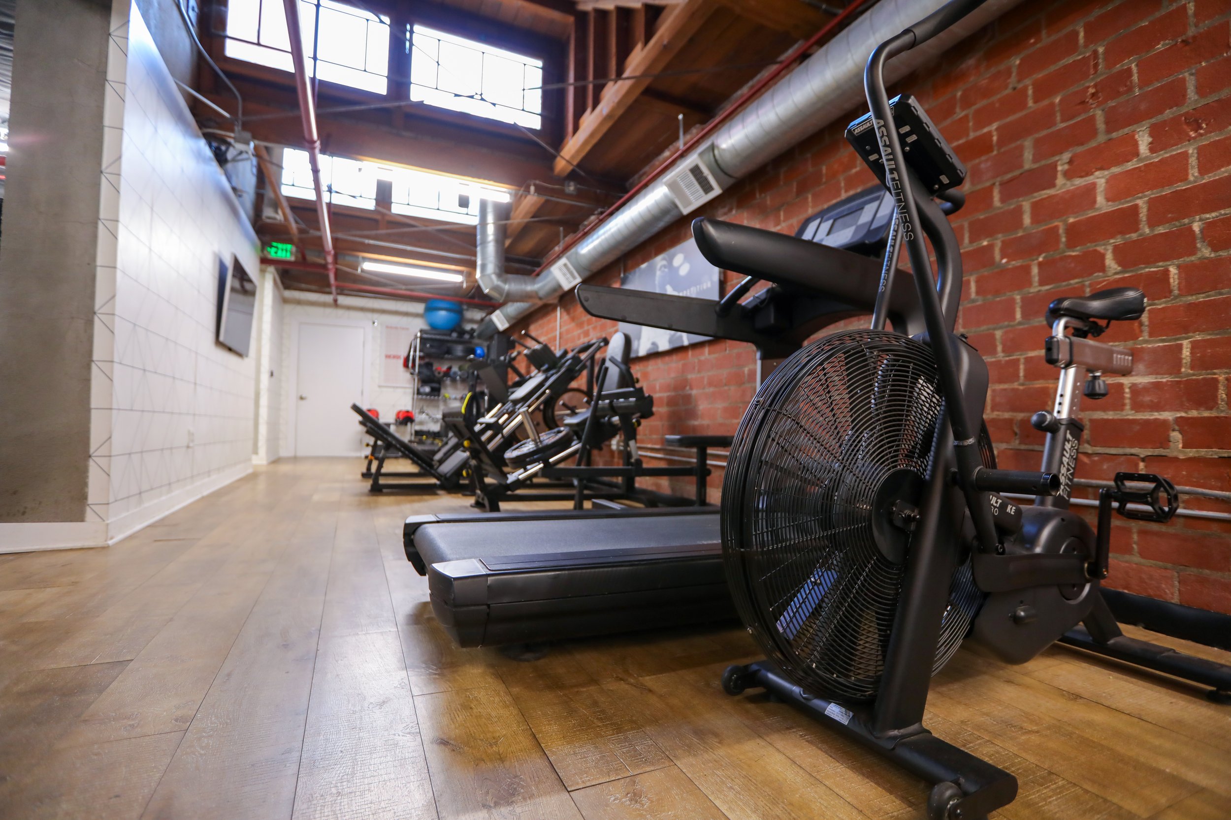 Dedicated cardio and lower body training corridor in a private Los Angeles fitness studio with treadmills, air bike, wood flooring, and natural light from skylights.