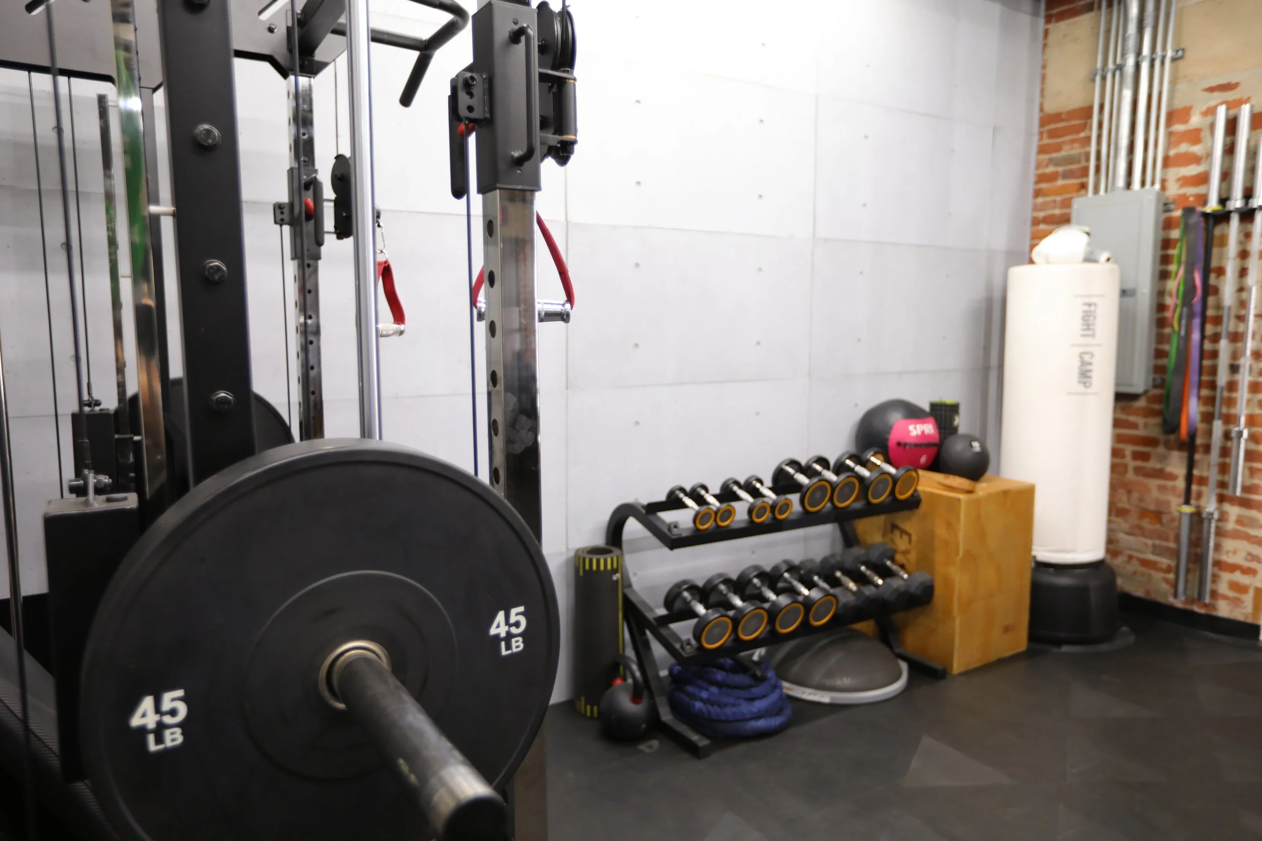 Gym workout area with a large 45-pound weight plate on a barbell, dumbbells on a rack, kettlebells, medicine balls, a foam roller, and resistance bands in a room with brick and concrete walls.