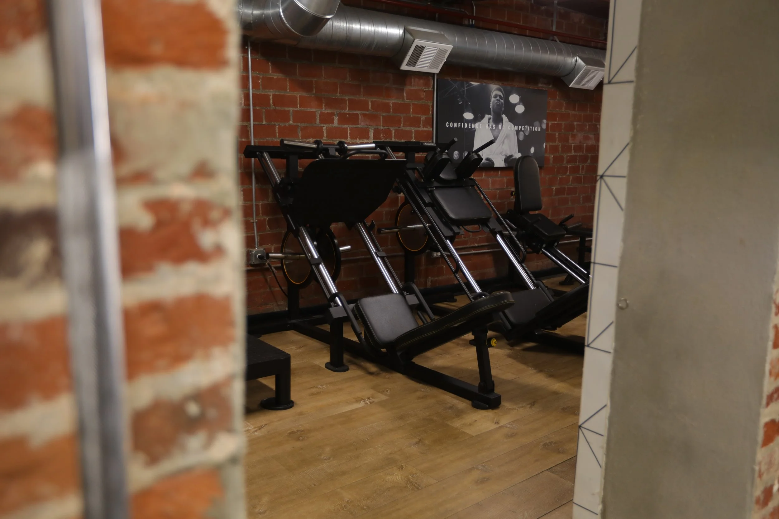 Gym space with black rowing machines against a brick wall, wooden floor, and a motivational poster of a person with the text "Confidence has no competition."