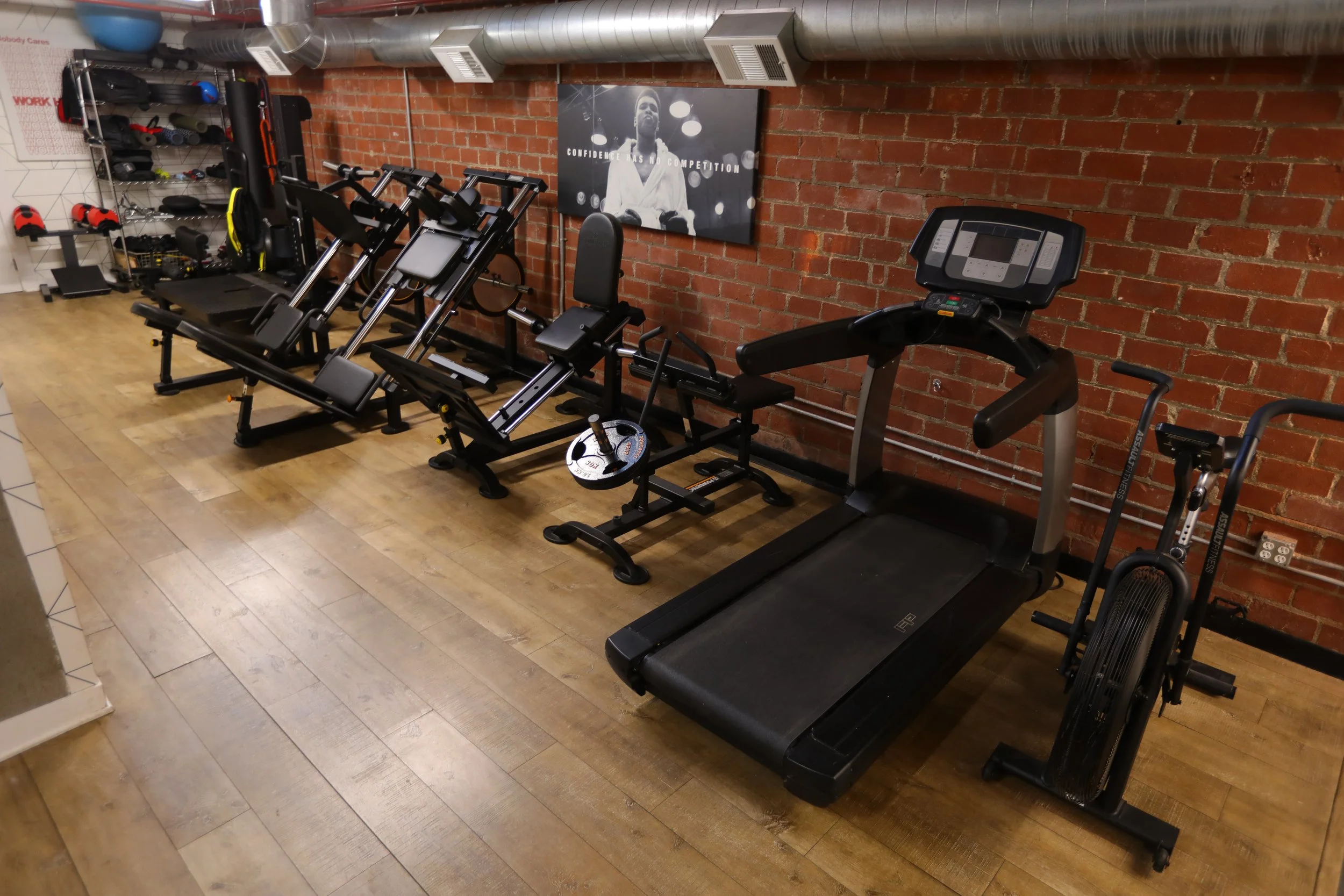 Lower body training area with leg press, treadmills, and cardio machines in a private Los Angeles fitness studio with exposed brick walls and wood flooring.
