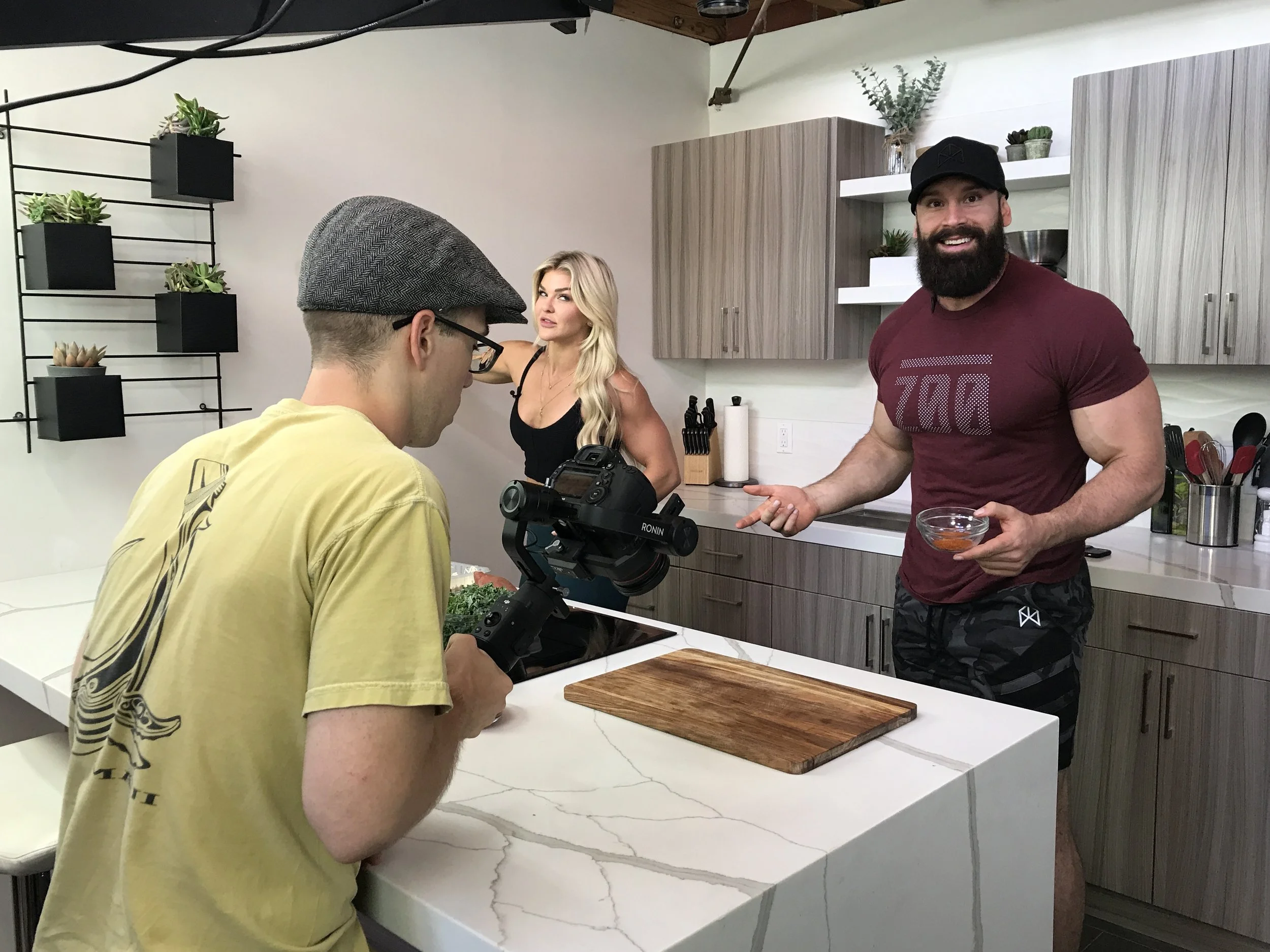 A man with a beard and a black cap, smiling, holds a small bowl of sauce while pointing at it. Two people are filming him in a modern kitchen, including a woman with long blonde hair and a man with glasses and a gray hat, who is operating a camera. The kitchen has wooden cabinets, white countertops, and decorative plant shelves.