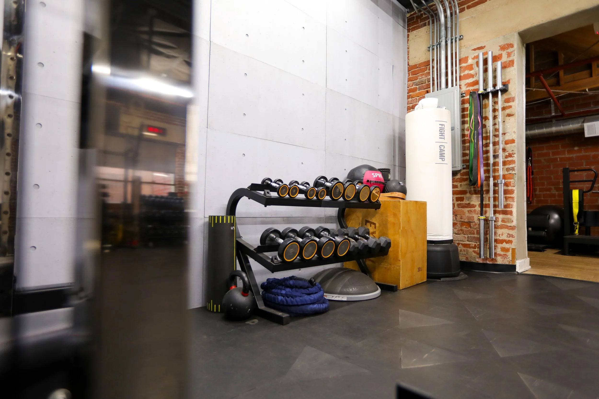 Interior view of a gym with dumbbells, kettlebells, medicine balls, a punching bag labeled 'FIGHT CAMP', resistance bands, and ceiling pipes against a concrete and brick wall.