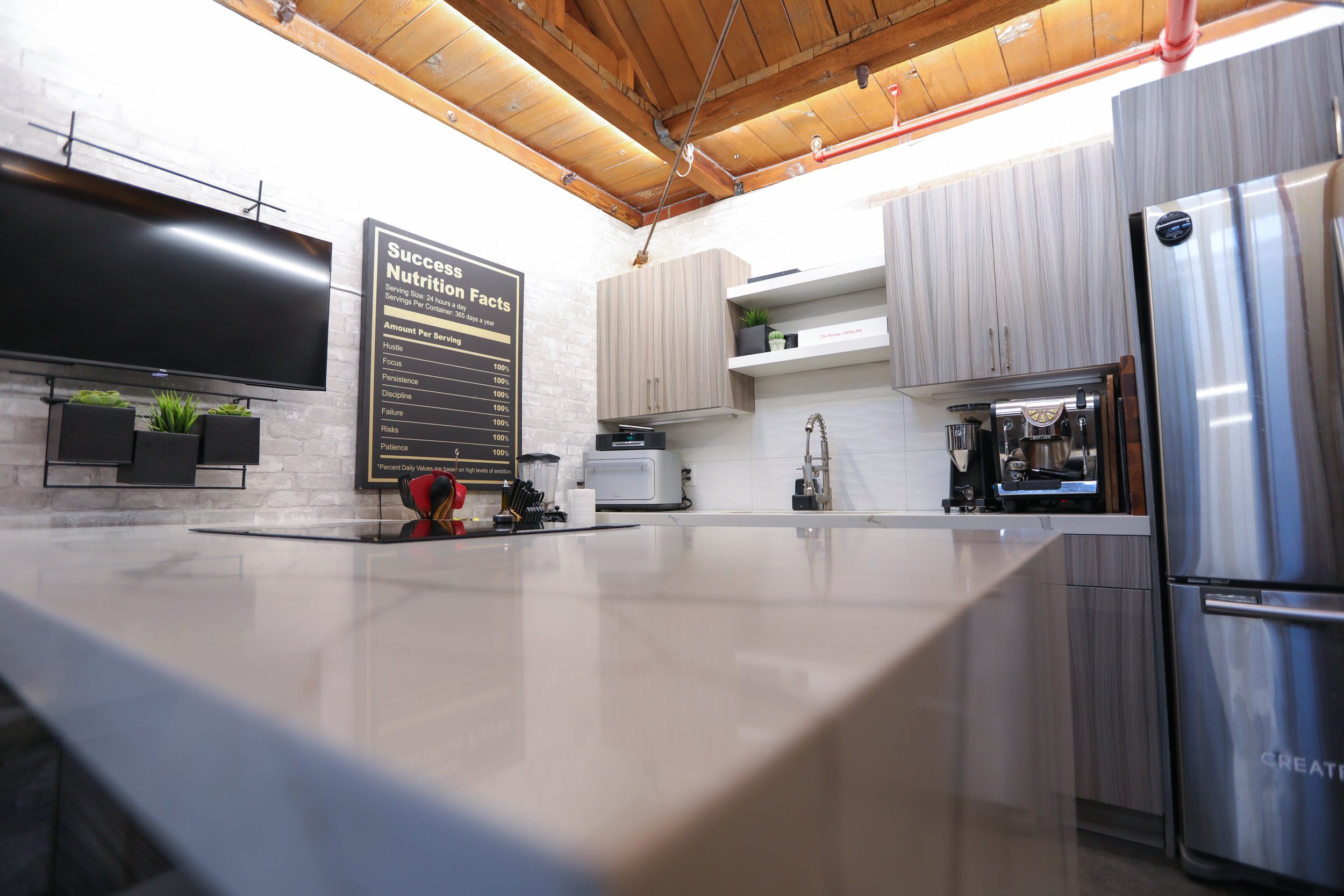 Modern kitchen with wood ceiling, gray cabinets, white brick wall, and stainless steel refrigerator. Small appliances and a blackboard with success nutrition facts are on the wall.