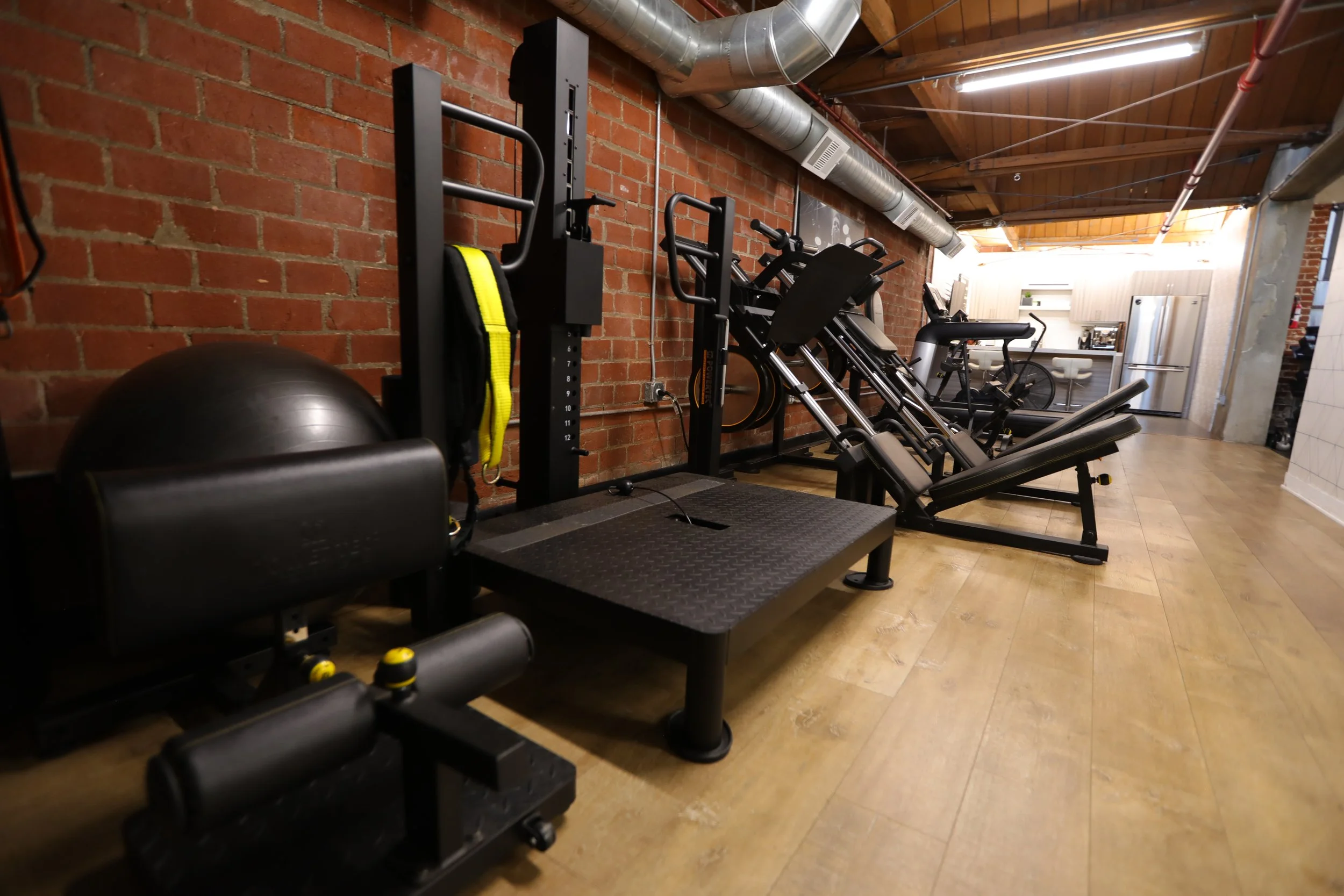 Lower body training corridor with leg machines, exposed brick walls, and wood flooring in a private Los Angeles fitness studio designed for personal training and content creation.