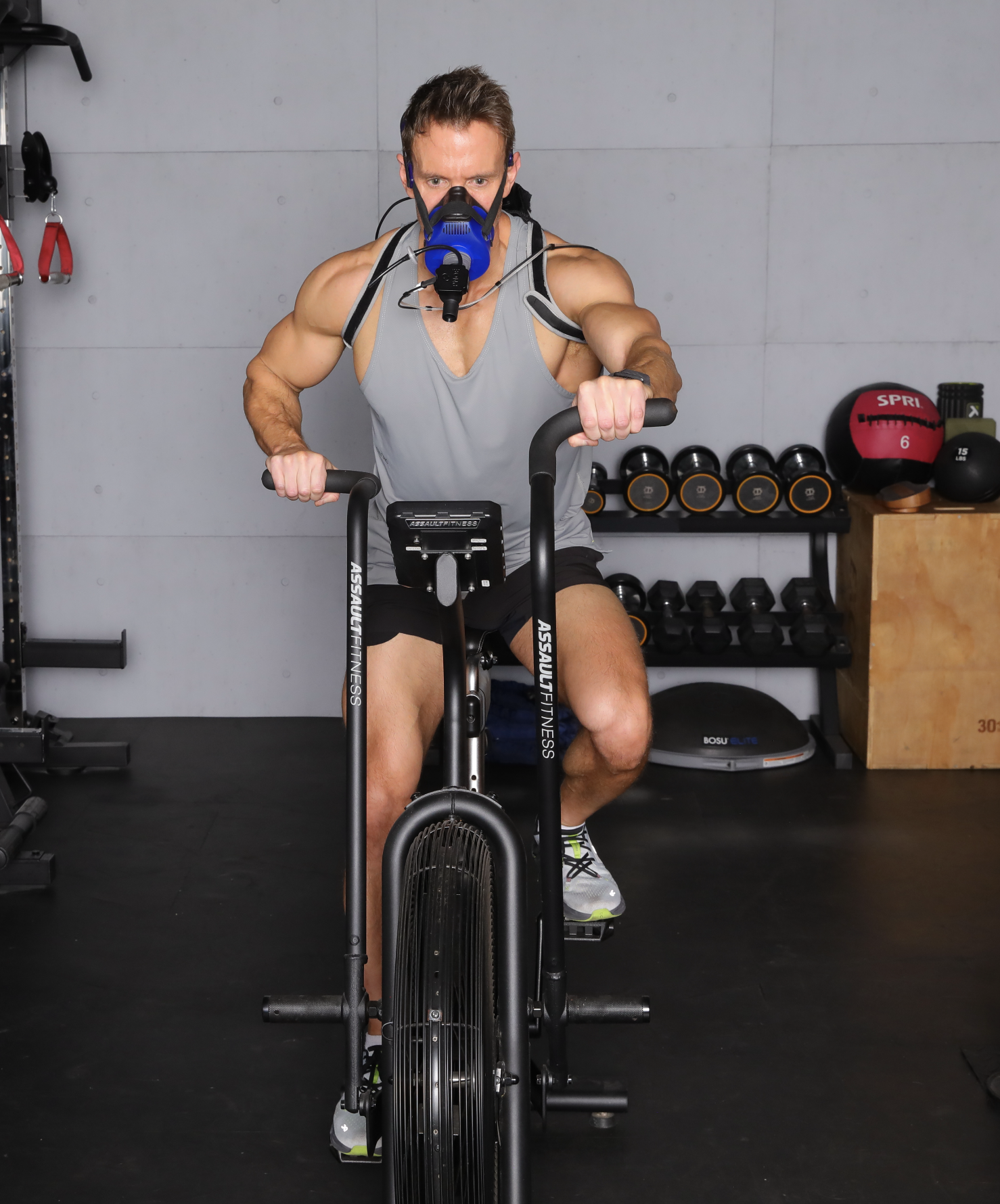 Man wearing a gray tank top, black shorts, and a breathing mask, riding a stationary exercise bike in a gym with black floor and workout equipment in the background.