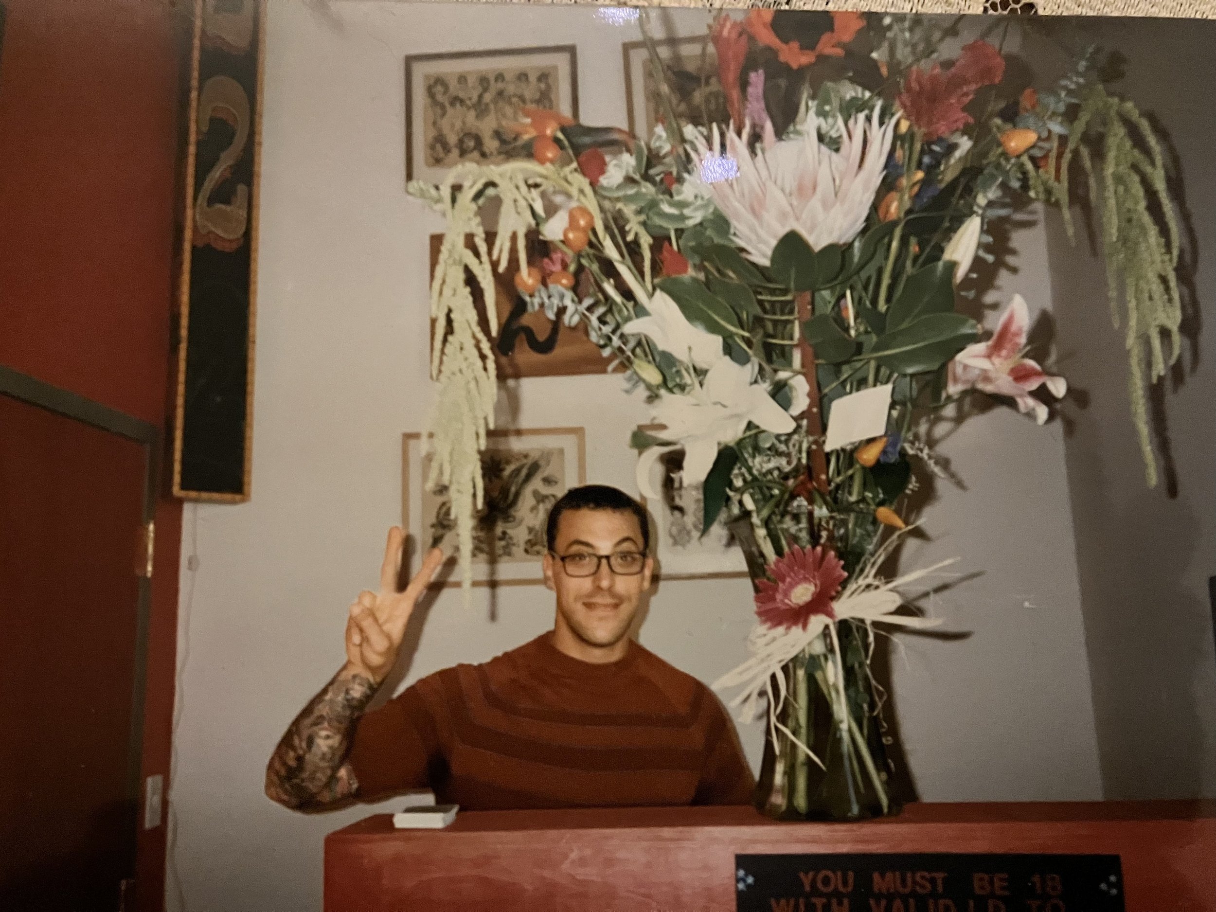 A person with glasses and a tattooed arm wearing a brown striped shirt, smiling and making a peace sign with their right hand, positioned behind a counter with a large floral arrangement in a vase. The background features framed pictures and a wall-mounted sign that reads 'You must be 18 with ID to be served'.