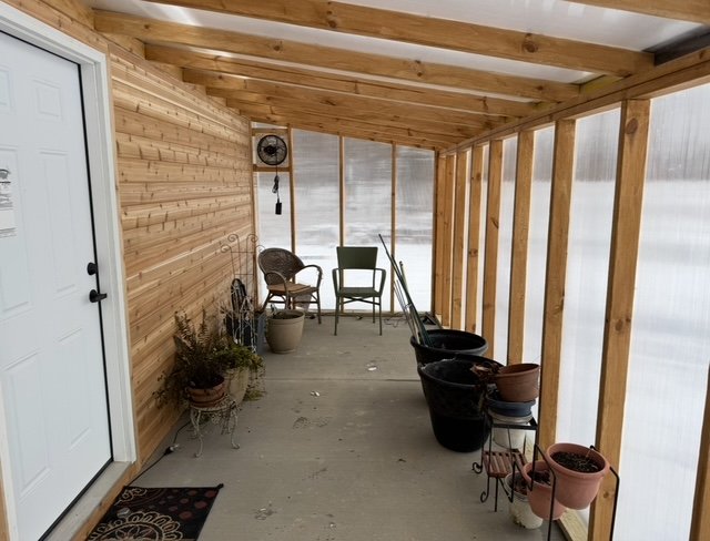 A covered porch with wooden walls and ceiling, several potted plants, two chairs, and empty buckets.
