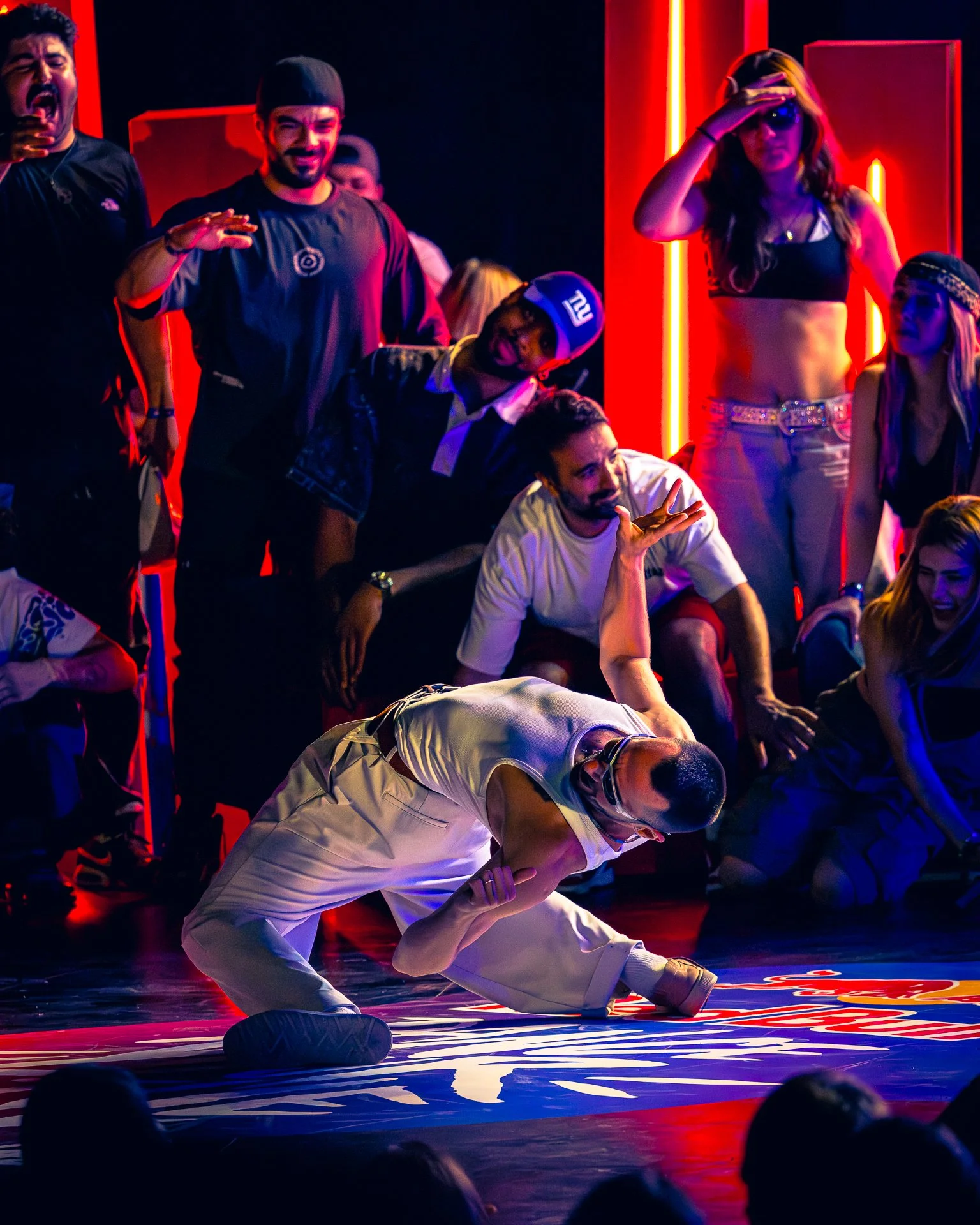 A breakdancer is performing a move on the floor surrounded by a group of onlookers. The scene is lit with colorful, neon lighting.