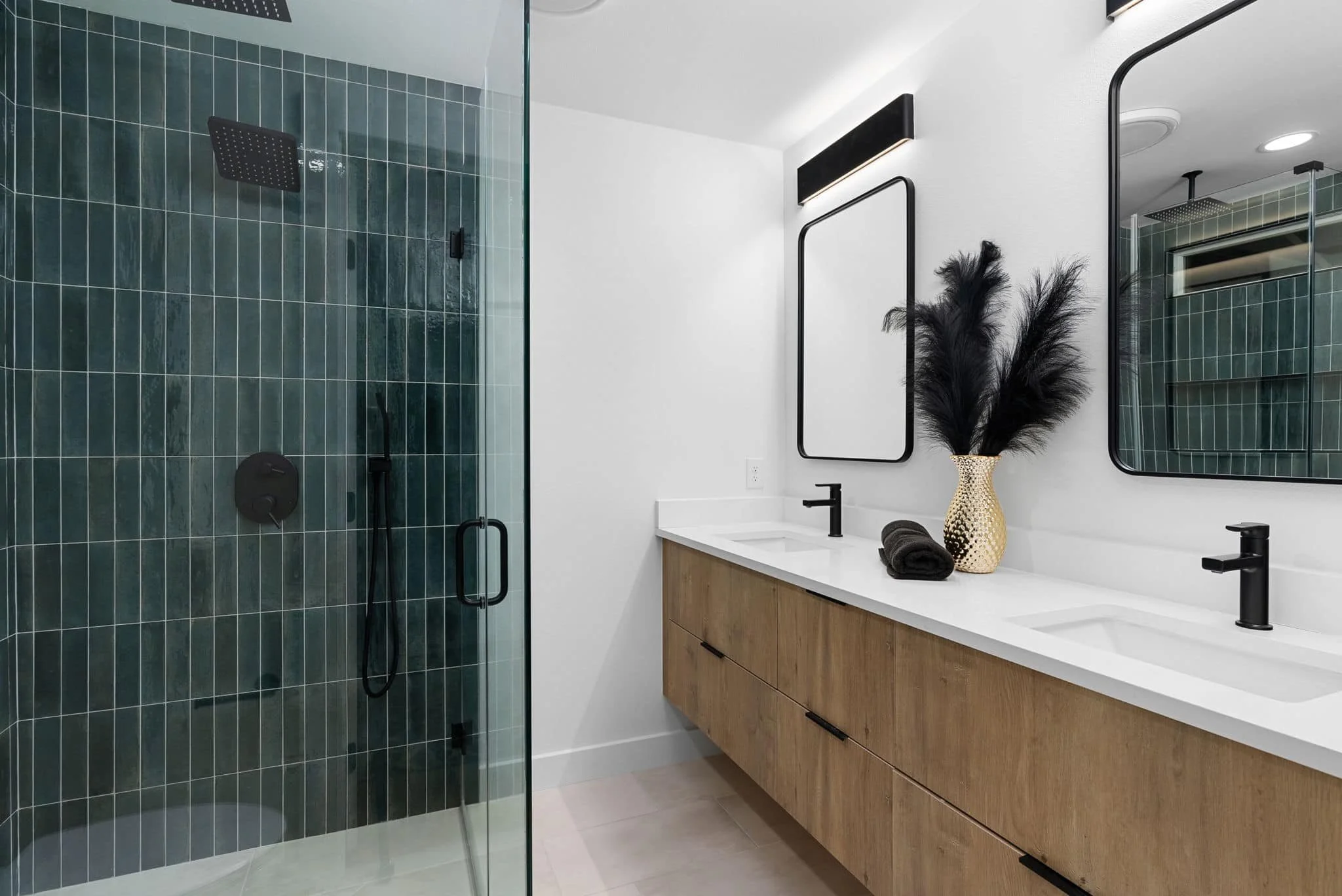 Modern bathroom with a glass walk-in shower with dark green tiles, a white vanity with two sinks, black faucets, a large mirror, and decorative black feathers in a gold vase.