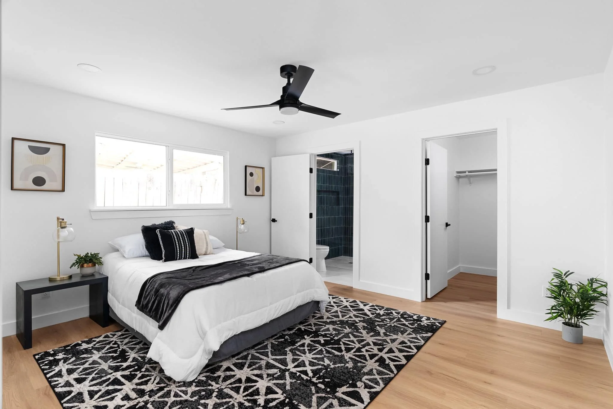 A modern, minimalistic bedroom with a white bed, black and white accents, a black ceiling fan, and an attached bathroom with a toilet. The room has light wood floors, a black and white geometric rug, and artwork on the walls. There's a window above the bed and a closet with an open door.