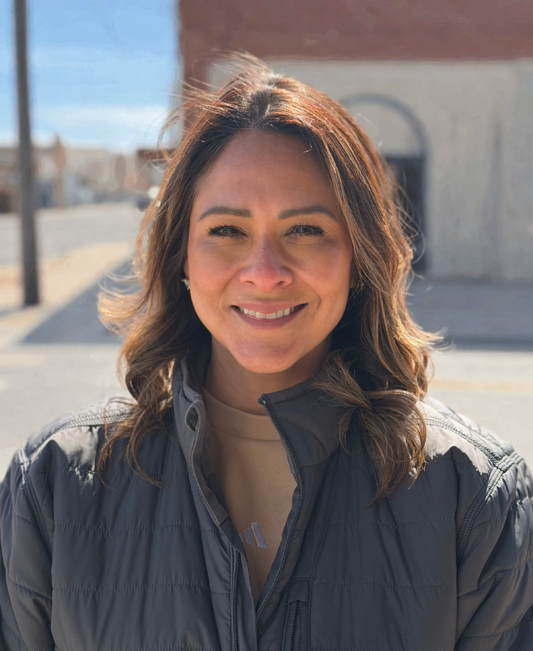 A woman smiling outdoors, wearing a gray puffer jacket and a cream-colored shirt, with sunlight on her brown hair. Background shows a building and a street under a clear blue sky.