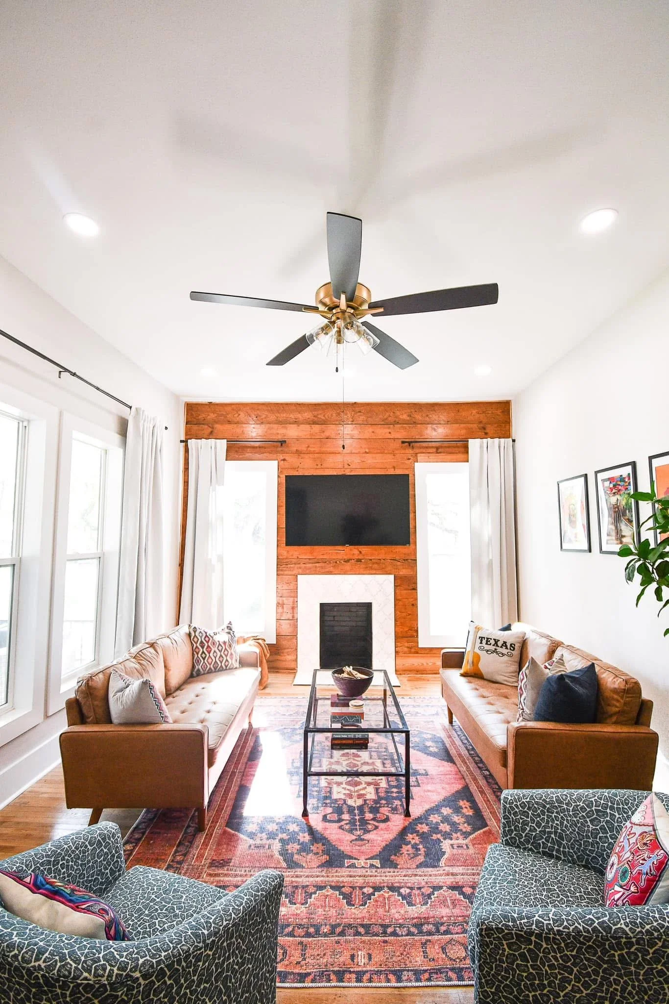 Living room with two leather sofas, a glass coffee table, patterned armchairs, a wood accent wall with a fireplace and a mounted television, large windows with white curtains, framed artwork on the wall, a ceiling fan with black blades, and a patterned area rug.